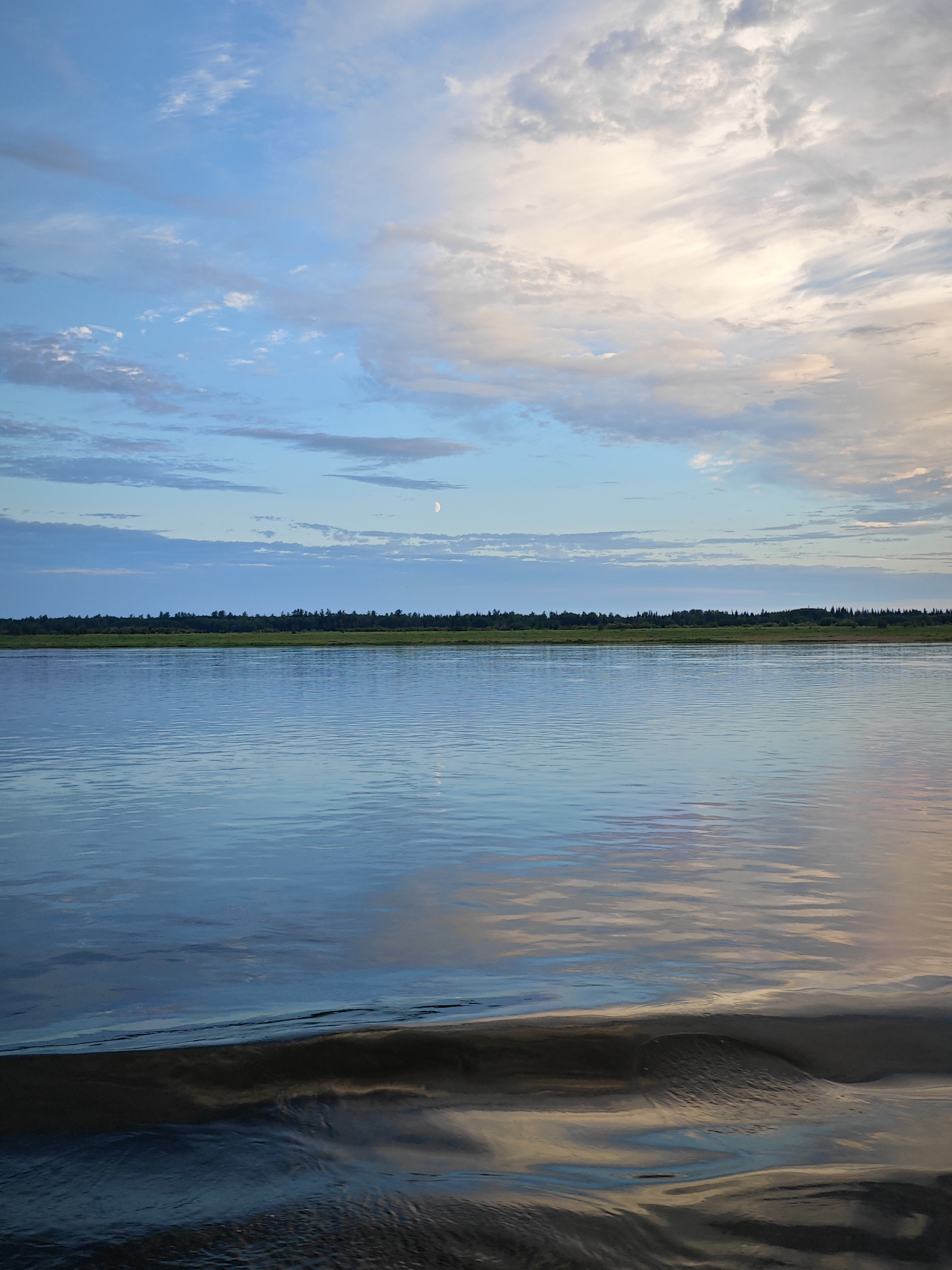This media file captures a serene landscape scene during twilight. The primary subjects are a calm body of water, a distant tree line, and a vast sky filled with clouds. The water occupies the foreground and mid-ground, reflecting the colors of the sky and appearing tranquil with gentle ripples. In the distance, a dense line of trees forms a dark horizon, suggesting a forested area. The sky above is a canvas of soft blues, whites, and hints of pink and orange, indicative of either sunrise or sunset. A crescent moon is faintly visible in the upper portion of the sky, further pinpointing the time of day.
The scene depicts a moment of natural beauty and quietude, with no human activity or man-made objects immediately apparent. The weather appears clear, with scattered clouds contributing to the picturesque sky. The overall mood is peaceful and contemplative. The location cues suggest a natural setting, possibly near a large river or lake, given the expanse of water and the distant forestation characteristic of the Nikulino, Russia region. The lighting and cloud formations suggest a peaceful transition between day and night. No visible text is present in the image.