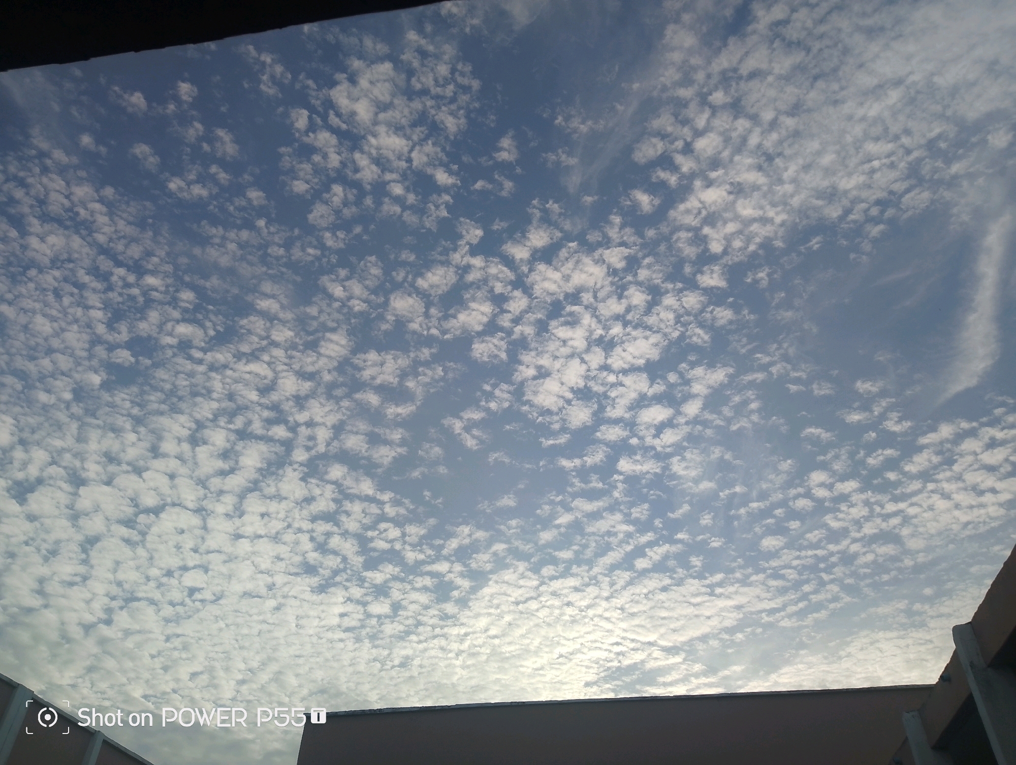 The image shows a view of a cloudy sky from below, looking upwards. The sky is a light blue with numerous small, white, scattered clouds that appear to be altocumulus clouds. A dark, horizontal band is visible at the top of the image, likely representing an overhang or structure blocking the view. At the bottom, the tops of buildings are visible, with a lighter colored structure in the foreground and a darker structure on the right. The text "Shot on POWER P55G" is visible in the lower left corner. There are no people or animals visible in the image. The scene depicts an outdoor environment, possibly urban, in Mairi, Nigeria.
