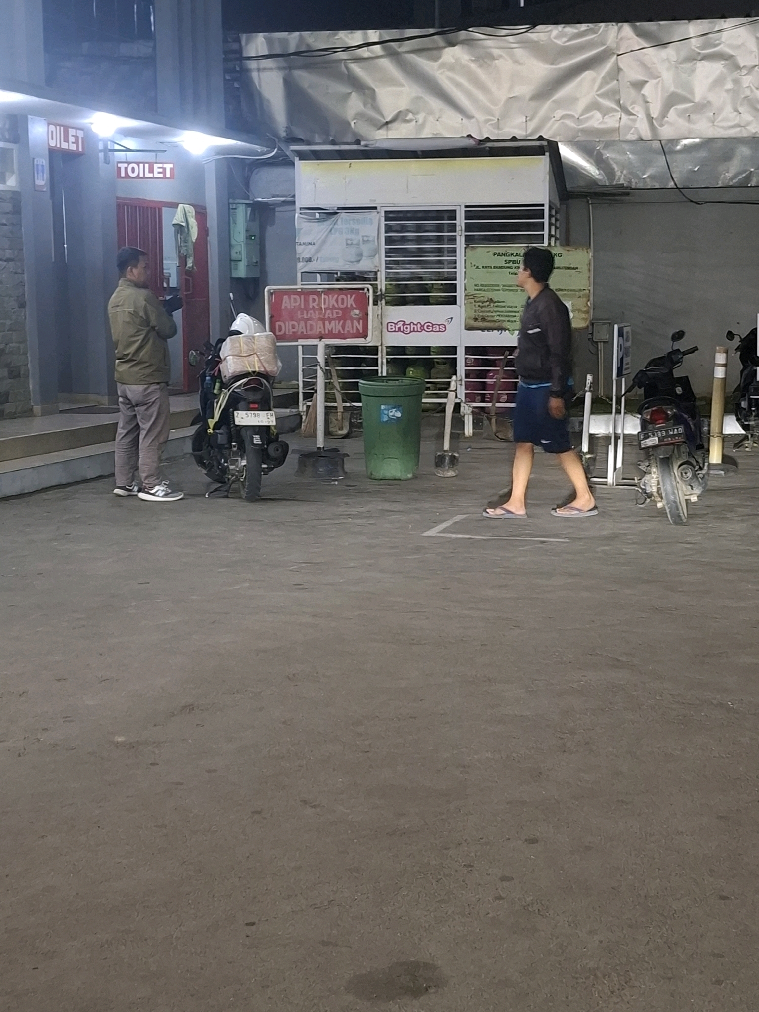 In Karangtengah, Indonesia, at what appears to be a gas station, two men are present. The man on the left stands near a building entrance with "Oilet" and "Toilet" signs illuminated in red. He is wearing a khaki jacket and grey pants, holding a dark object in his hands, possibly a phone. To his right, a motorcycle is parked, loaded with packages wrapped in brown paper and secured with yellow straps. The man on the right is walking away from the camera, wearing a dark jacket and blue shorts, with sandals on his feet. A second motorcycle is parked to his right. Behind the men, a structure with shelves holds various items, including gas tanks, and a sign for "Bright Gas" is visible. Another sign, in red, reads "API ROKOK HARAP DIPADAMKAN," which translates to "Fire cigarettes please extinguish." A green trash can is located to the left of the gas station structure. The ground is paved with asphalt, and white lines mark a designated area. The lighting suggests it is nighttime or dusk.