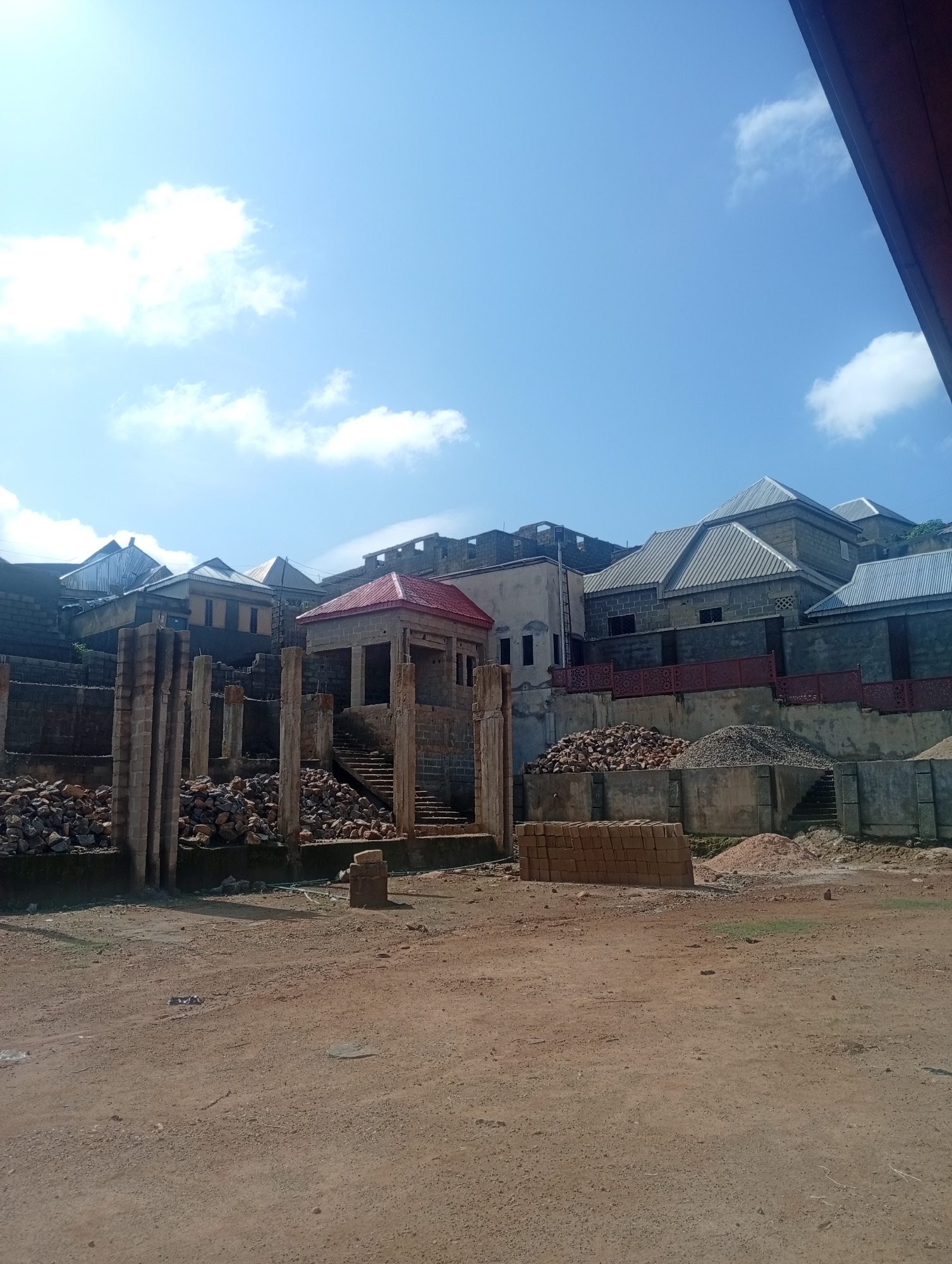 In Jos, Nigeria, a construction site is depicted under a clear blue sky with scattered white clouds. The scene is dominated by various stages of building development, suggesting a residential area under construction. In the foreground, a dirt ground is visible, with a neat stack of red bricks on the right side. Behind the bricks, a pile of sand and gravel are situated next to a concrete wall, which is adorned with a decorative metal railing. Further back, a partially constructed building with concrete pillars and exposed brickwork stands out. This structure features a red-tiled roof and a set of stairs leading upwards. Beyond this, a row of finished houses with corrugated metal roofs and stone walls are visible on a higher elevation. The overall impression is of a developing neighborhood in Jos, with a mix of ongoing construction and completed structures.