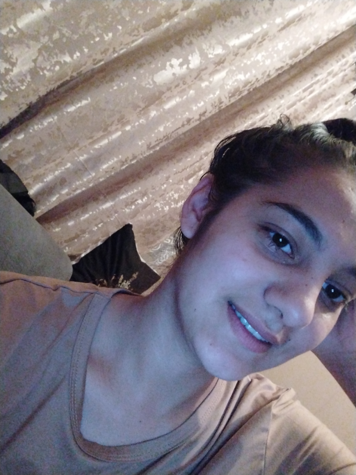 A close-up selfie of a person with dark hair pulled back, fair skin, and visible teeth. The person is wearing a brown top. The background consists of a tan, textured curtain. A dark pillow is partially visible on the left side of the frame, along with a gray couch. The location is Unknown, Unknown.