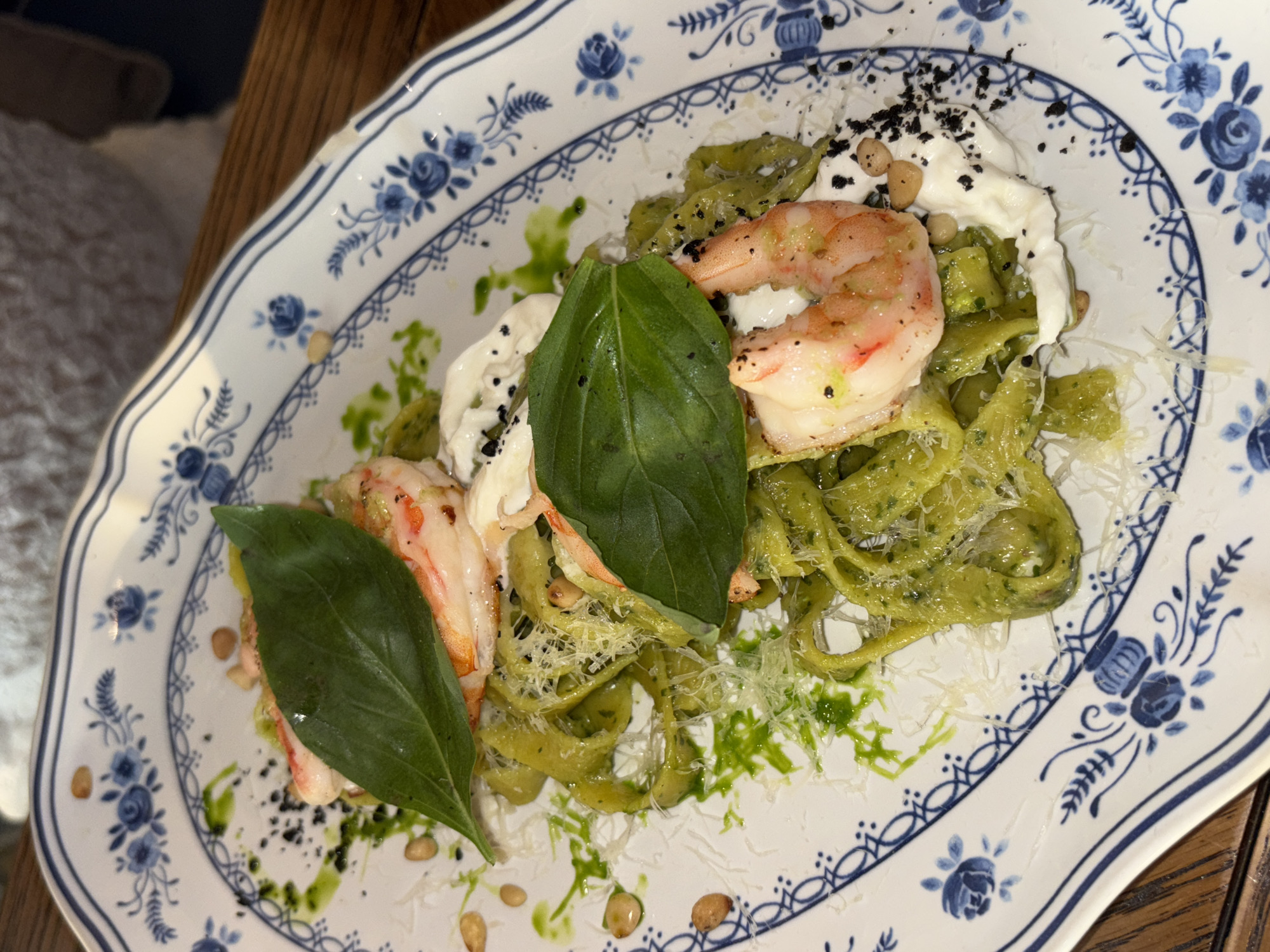 A meticulously plated dish is centrally featured on a white ceramic platter with an intricate blue floral and geometric border pattern. The main course consists of green ribbon pasta, likely tagliatelle or pappardelle, generously coated in a vivid green sauce, possibly pesto. Four cooked, peeled shrimp, exhibiting a pinkish-orange hue, are strategically placed atop the pasta. Two prominent, large green basil leaves serve as a garnish, enhancing the dish's visual appeal. Portions of a creamy white ingredient, resembling burrata or a similar soft cheese, are distributed among the pasta and shrimp. The dish is further embellished with scattered pine nuts and finely grated white cheese. Dark, granulated specks, potentially black pepper or a decorative spice, are sprinkled over the creamy elements. A thin, bright green liquid or oil is artfully drizzled across the plate and food. The plate is positioned on a dark wooden surface, visible to the right, with a light-colored, textured fabric element visible in the upper left background. The scene is located in Perm, Russia.