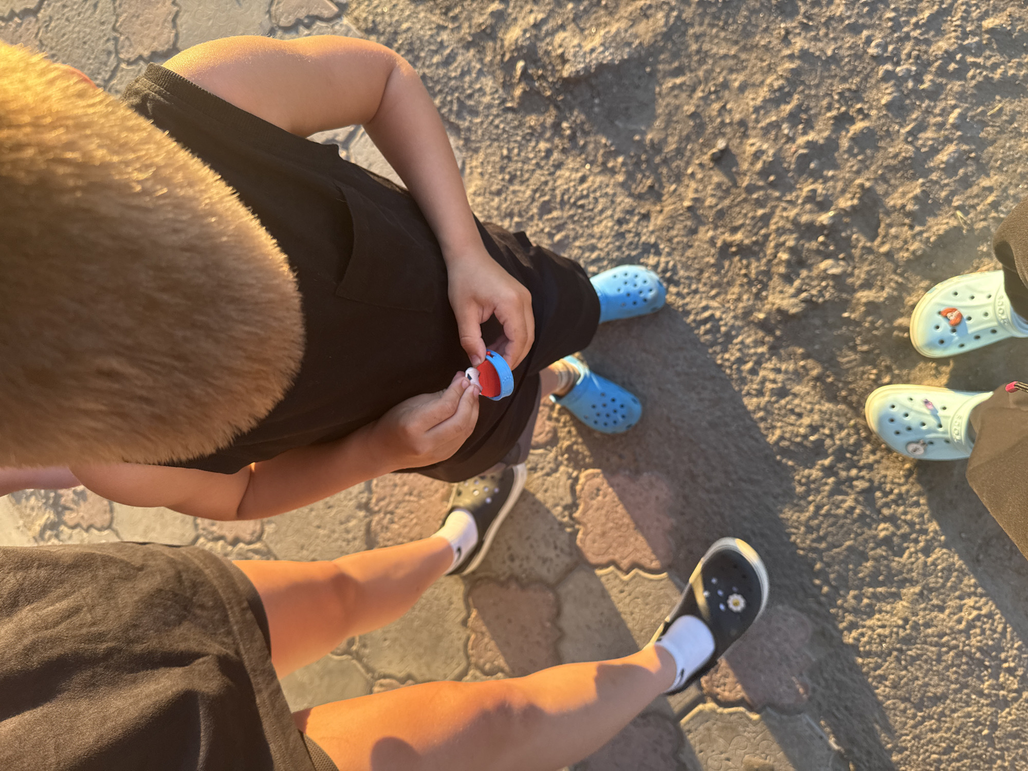 The image shows a close-up view of the lower legs and feet of three individuals standing on a paved surface with patches of dirt or sand. The lighting suggests it is late afternoon or early evening, with strong shadows cast on the ground.
One person, seen from the waist down, is wearing a dark skirt and black slip-on shoes with white socks. The shoes have a daisy-like embellishment. Their legs are illuminated by golden light.
A child, also seen from the torso down, is wearing a black sleeveless shirt and bright blue Crocs. The child is holding a small red and blue toy in their hands, which are behind their back. The child's arms and hair are visible.
Another person's feet, wearing light blue Crocs with a red embellishment, are visible at the bottom of the frame. They are wearing dark pants.
The ground is a mix of light-colored pavers and rough, sandy soil. The scene appears to be outdoors, possibly in a park or walkway area. There is no visual information in the image to place the location specifically in Dnipro, Ukraine.