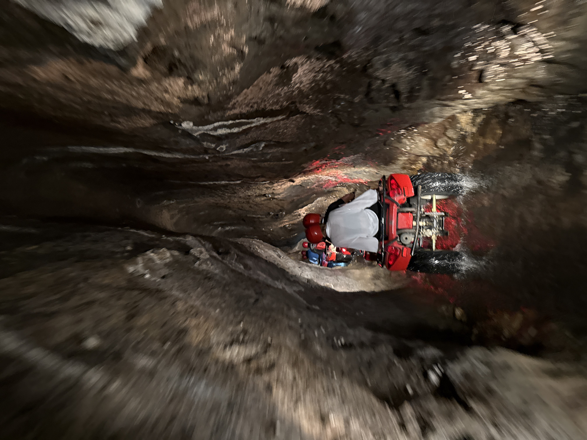 A group of individuals on red All-Terrain Vehicles (ATVs) navigates a narrow, wet, and dimly lit natural cave or tunnel in Payangan, Indonesia. The perspective is from behind one ATV, looking forward as a procession moves through the subterranean passage. The cave walls are rugged, textured, and appear damp or wet, with areas reflecting light from the vehicles. Shallow water covers the cave floor, and spray is visible from the tires of the moving ATVs. Each rider wears a red helmet; the closest rider is dressed in a white long-sleeved shirt. Red taillights from the ATVs illuminate the water and the immediate surroundings, casting a reddish hue on the lower cave walls. Several more ATVs and riders are visible in a single file ahead, disappearing into the darker interior of the cave. The scene depicts an organized recreational adventure activity.