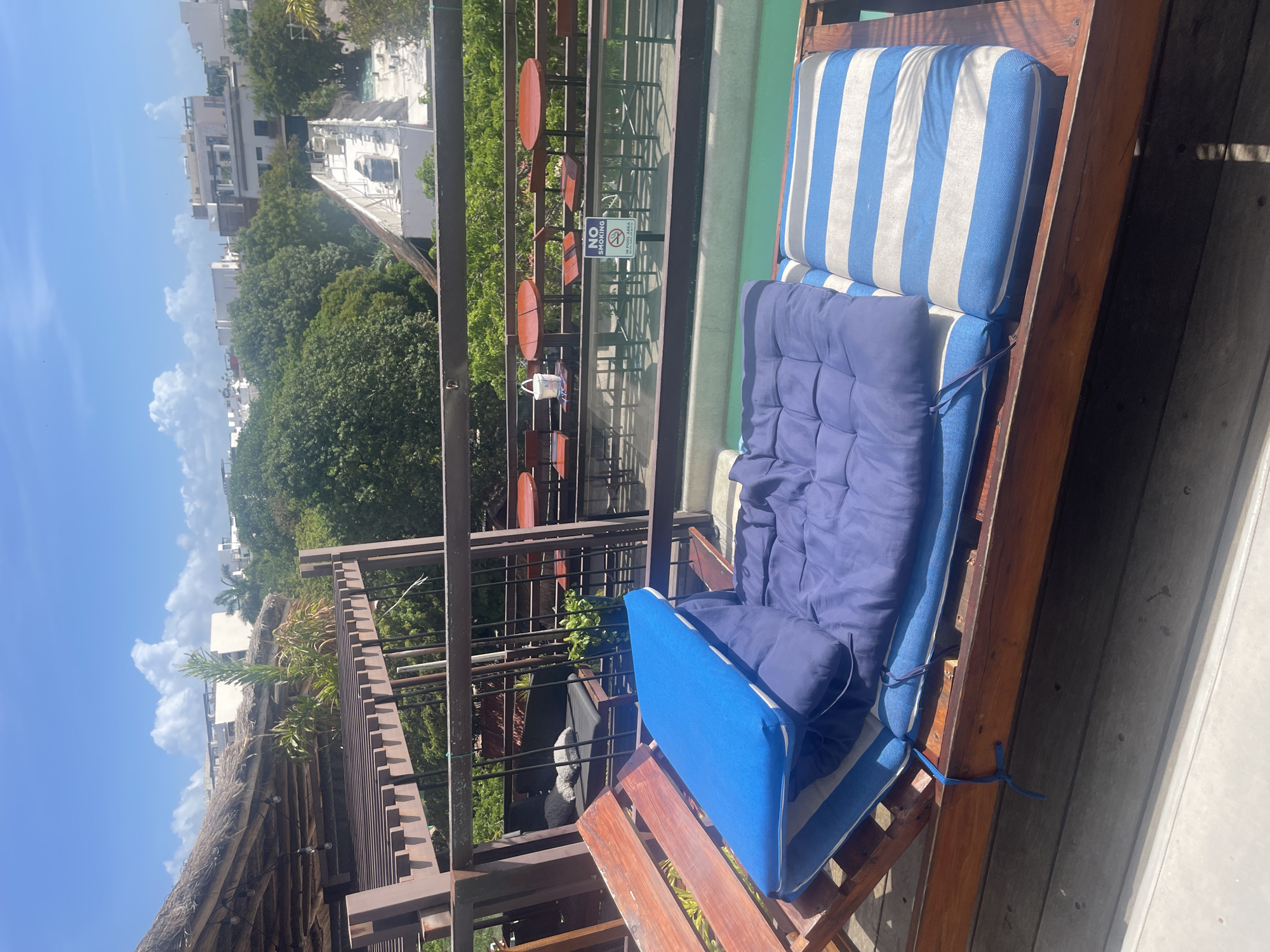 The image shows a rooftop setting in Playa del Carmen, Mexico, indicated by the architecture and vegetation in the background. A wooden patio lounge chair with blue and white striped cushions and additional solid blue cushions is in the foreground. The lounge chair is situated on a wooden deck. Beyond the deck, a metal railing overlooks the urban landscape with buildings and green trees. A "No Smoking" sign is visible near outdoor tables and chairs on a terrace. The sky is blue with some clouds.