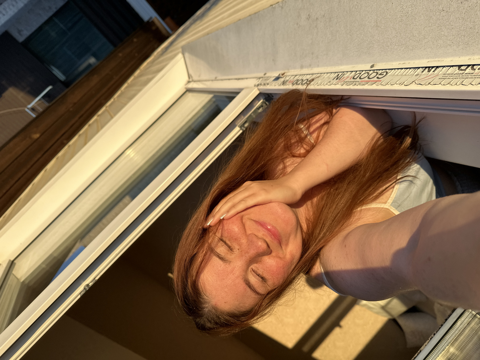A young woman with long, auburn hair and freckles is leaning out of a window, her eyes closed and a gentle smile on her face. The setting sun casts a warm, golden light on her skin, highlighting her features. She is resting her right cheek on her hand. The background shows the interior of a room and the exterior of a building with wooden and white siding. The image appears to be taken in Nizhny Novgorod, Russia, based on the context provided.