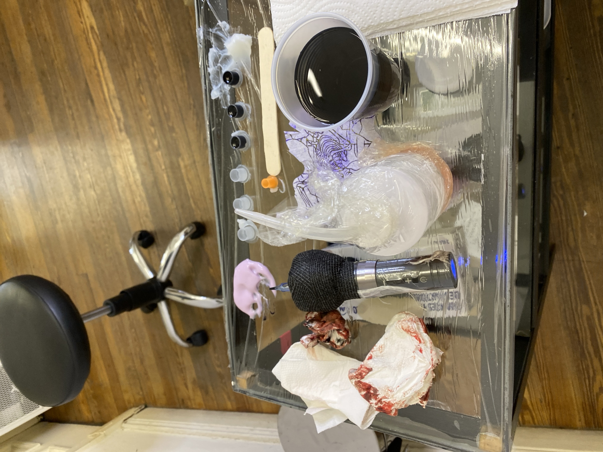 The image depicts a close-up of a tattoo artist's workstation, viewed from above. The workstation is a clear, likely plexiglass, surface covered in plastic wrap for sanitation. The scene contains various tattoo-related tools and supplies. There's a cup of black tattoo ink, several small ink cups, a wooden stirring stick, and a tattoo machine wrapped with a textured black grip and blue light. Also visible are bloody tissues and a dollop of pink ointment. Under the workstation is part of a chair. The location is unknown, and there's no information indicating it's in Buenos Aires, Argentina. The scene suggests a tattoo in progress or recently completed. There is no visible person in the image.