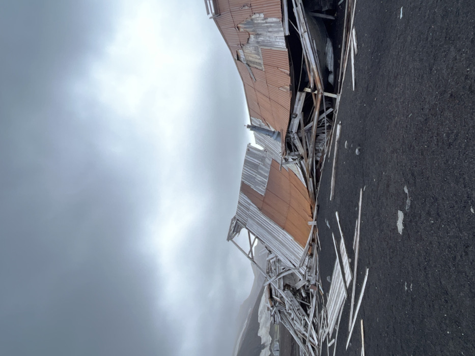 A dilapidated building with rusted corrugated metal siding is partially collapsed, exposing its wooden frame. The structure is situated on dark, volcanic-looking terrain under an overcast sky. Debris from the building is scattered on the ground. The overall scene suggests decay and abandonment in a harsh environment.