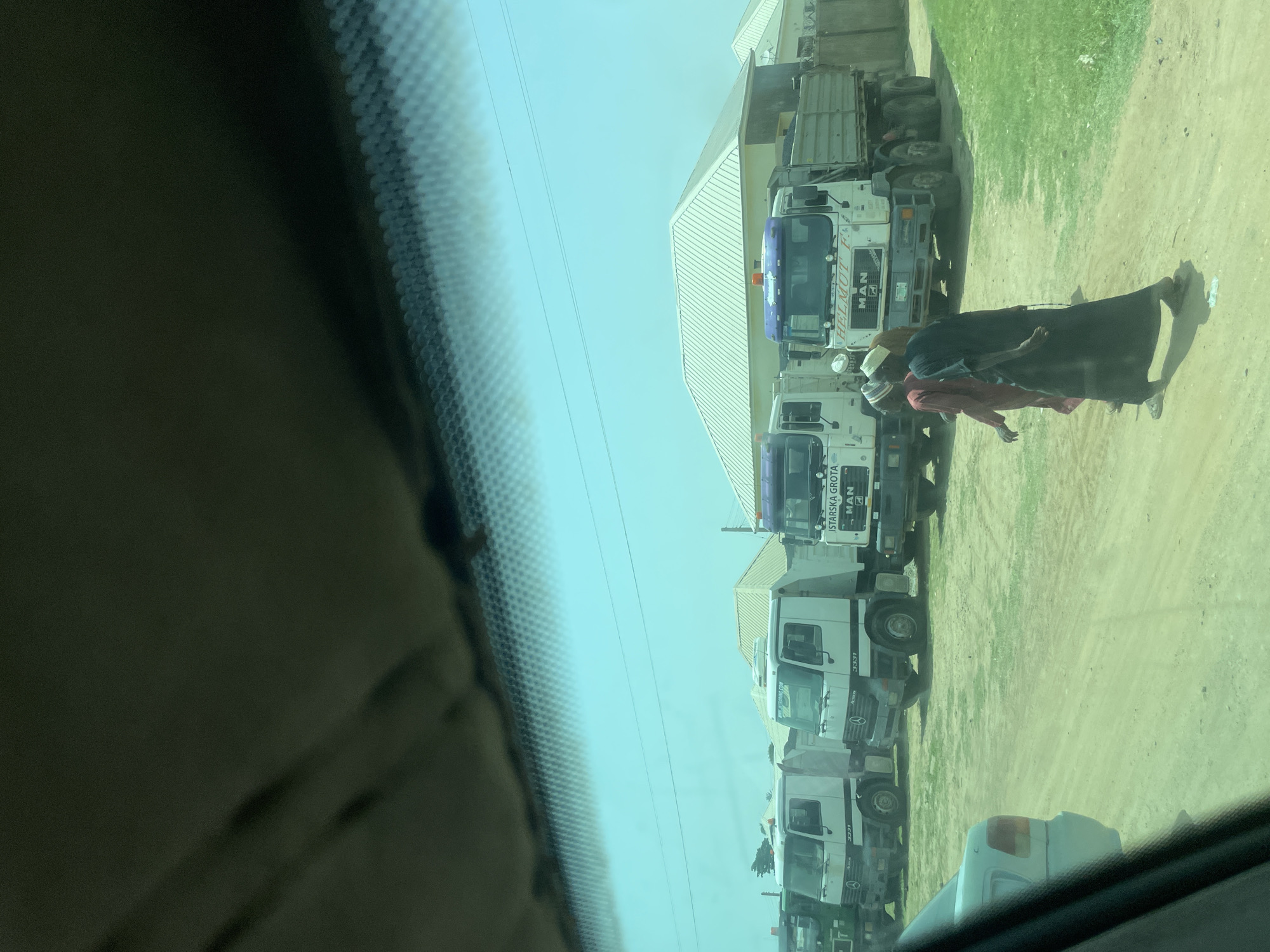 The image shows a line of white trucks parked on a dirt and grass area, possibly a lot or service area. The trucks are of various models, with some displaying "MAN" branding. One truck has a purple stripe on its cab. In the foreground, a person wearing a dark blue or black robe and a light-colored headscarf is visible, along with another person in a reddish garment, also with a head covering. They are walking or standing near one of the trucks. The sky is bright and appears clear, suggesting daytime with fair weather. The overall scene seems to be outdoors in a relatively undeveloped area, given the dirt road and sparse vegetation. The image is taken from inside a vehicle, with a portion of the windshield and dashboard visible in the bottom and left foreground. Text visible on the trucks includes "MAN", "3331", "HELMUT F.", and "STVARSKA GROTA". The setting appears to be Bauchi, Nigeria. The activity is not entirely clear, but it involves people interacting with or in the vicinity of parked trucks.