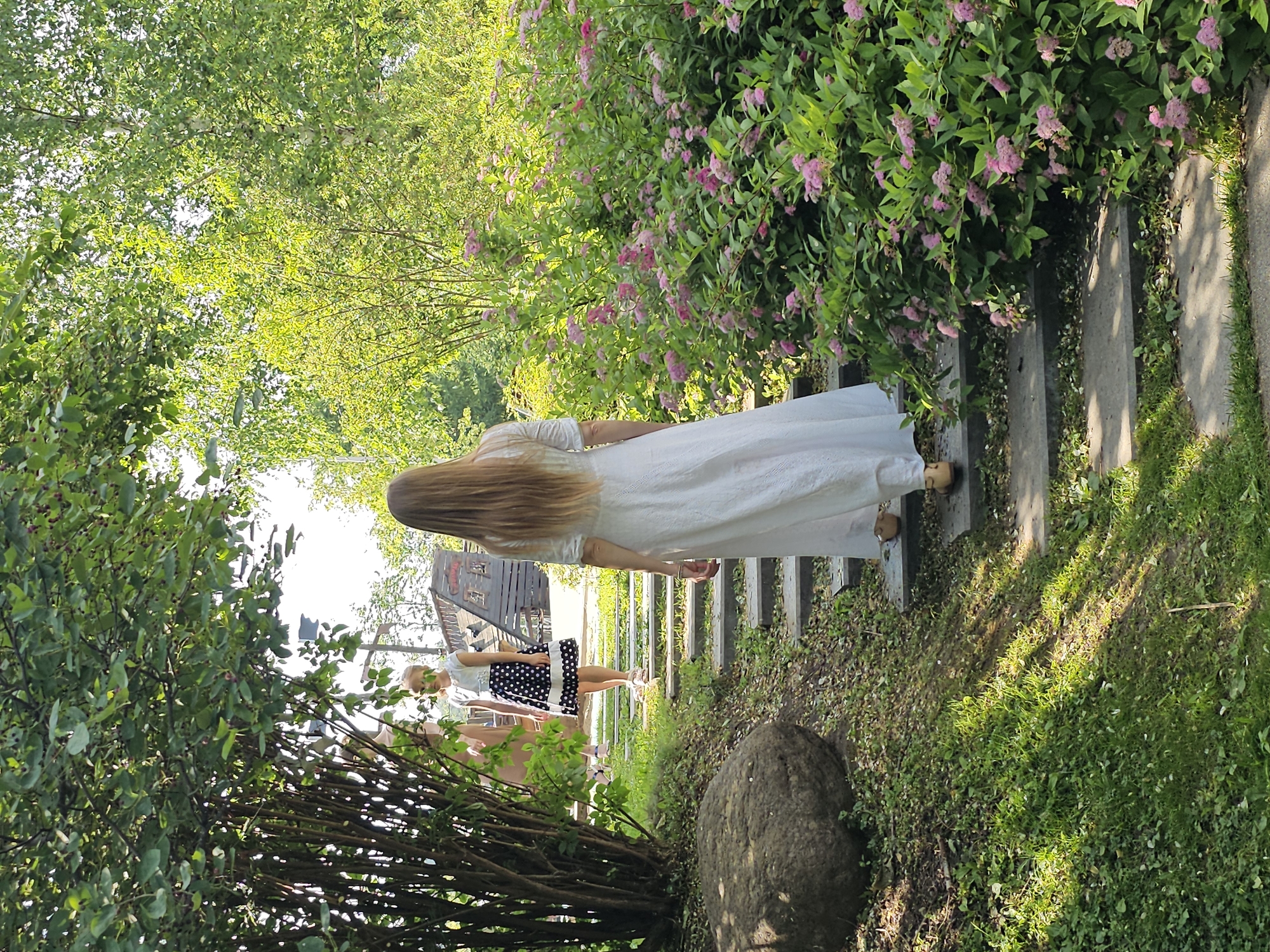 The image depicts two individuals in an outdoor setting, likely a park or garden. In the foreground, a person with long, blonde hair is walking up a set of stone steps, facing away from the camera. They are wearing a long, white, eyelet lace dress and beige flat shoes. To the left of the steps and slightly behind the person in white, another individual, a young girl, is visible, also walking up the steps. She has blonde hair and is wearing a white top with short sleeves and a black and white polka dot skirt. Behind them, a wooden structure, possibly a playground or playhouse, is partially visible. The environment is lush and green, with trees providing shade and a dense bush with small pink flowers to the right of the steps. The ground is covered in grass and fallen leaves, and a large, round rock is situated to the left of the steps. The scene is illuminated by bright sunlight, casting shadows on the grass and steps.