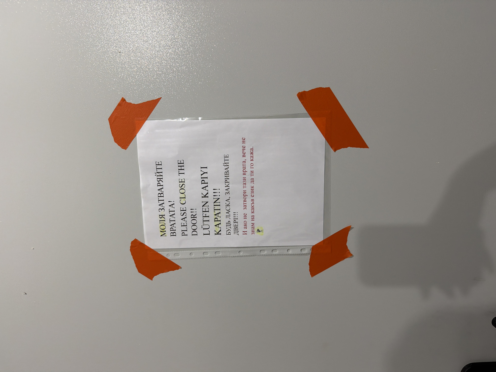 The media file displays a piece of paper taped to a plain, textured, light-colored wall. The paper contains text in multiple languages, including Bulgarian, English, Turkish, and Russian. The prominent message, repeated in different languages, is "PLEASE CLOSE THE DOOR!!". The text also includes phrases like "МОЛЯ ЗАТВАРЯЙТЕ ВРАТАТА!", "LÜTFEN KAPIYI KAPATIN!!!", "БУДЬ ЛАСКА, ЗАКРИВАЙТЕ ДВЕРІ!!!", and a humorous closing in Bulgarian: "И ако не затвори тази врата, вече не знам на какъв език да ти го кажа." (And if you don't close this door, I don't know in which language to tell you anymore.)
The paper is laminated and held to the wall with four pieces of orange tape, one at each corner, giving it a slightly crumpled appearance. The lighting suggests an indoor setting, possibly during daylight hours, casting faint shadows on the wall. There are no people visible in the image, and the focus is entirely on the informational sign. The overall impression is a practical, perhaps slightly quirky, instruction placed in a communal or shared space to remind people to close the door. The Plovdiv, Bulgaria location context implies this sign might be found in a building or public space within that city.
