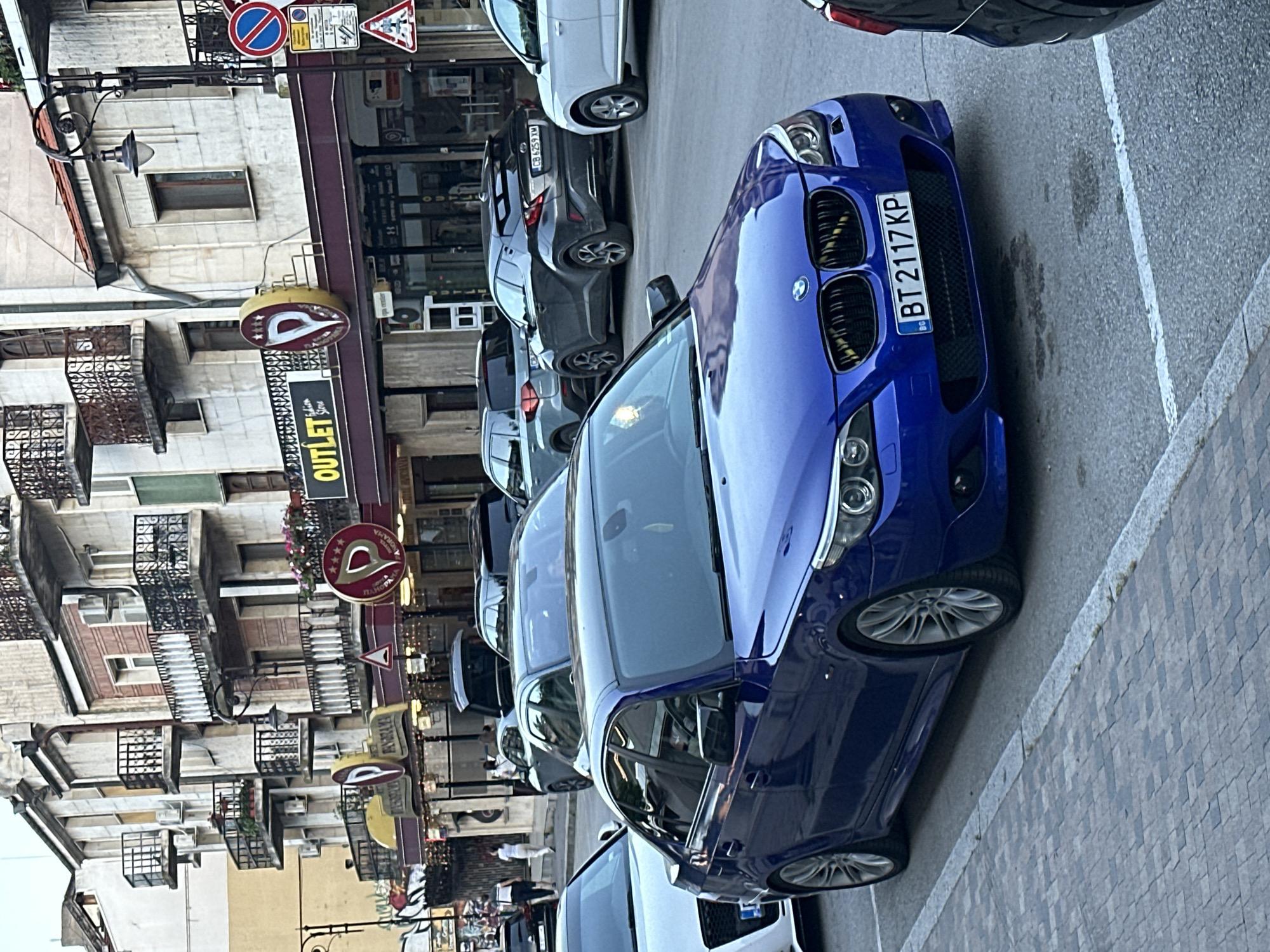 A blue BMW sedan with license plate BT 2117 KP is parked on a street in Veliko Tarnovo, Bulgaria. The car is in the foreground, facing right. Behind it, several other cars are parked, including a white Audi to its left and a dark gray Toyota C-HR with license plate CB 6259 XM to its right. In the background, a multi-story building with balconies and shops on the ground floor is visible. One of the shops has a sign that reads "OUTLET Store" and another with a red circular logo featuring a stylized "P" and stars. Several pedestrians are walking on the sidewalk in front of the shops. The street is paved with cobblestones. The overall scene depicts a street view in a European city.