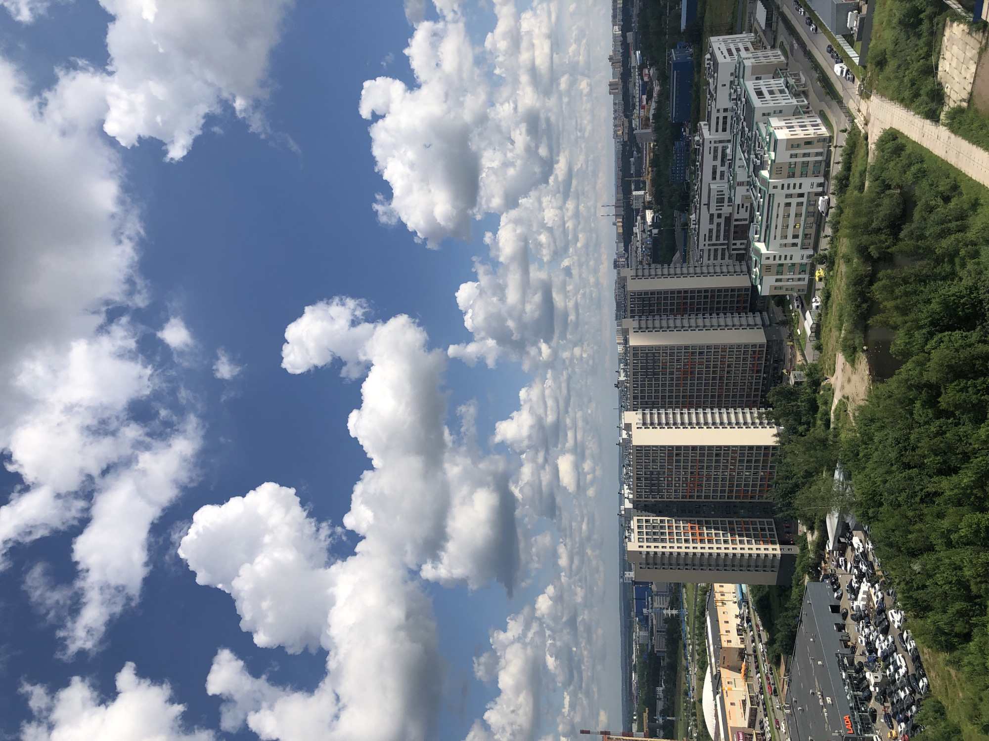 An aerial view captures a cityscape under a bright blue sky dotted with fluffy white clouds. The foreground is dominated by lush green trees, some of which appear to be bordering a small body of water. To the left, a large parking lot is filled with numerous cars and vans, adjacent to a building with a prominent "LADA" sign. Further into the scene, several tall, modern residential buildings with light-colored facades and orange window accents stand prominently. Beyond these, a collection of lower, contemporary buildings with white exteriors and green roofs are visible, along with other urban structures and industrial buildings in the distance. The overall impression is of a developing urban environment with a mix of residential, commercial, and industrial areas.