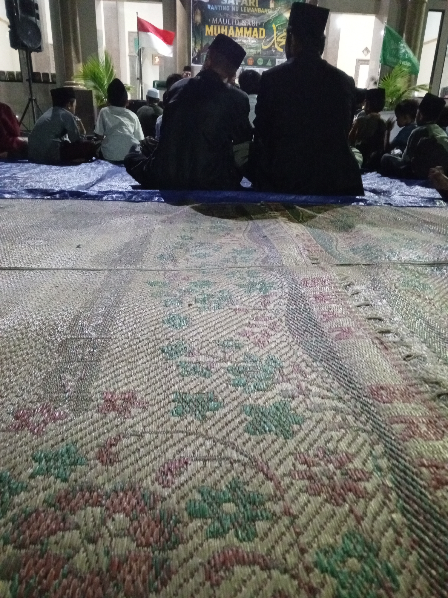 In Lemahbang, Indonesia, a group of individuals, primarily young males, are gathered indoors, seated on a patterned woven mat. The setting appears to be a communal gathering or event, possibly a religious observance, as indicated by the Islamic attire of some individuals, including peci hats. In the background, a banner with text, partially visible and likely in Indonesian, suggests an event related to "MAULID NABI MUHAMMAD." A red and white Indonesian flag is displayed prominently, alongside a green flag with Arabic script. A speaker on a stand is also visible. The lighting is dim, suggesting an evening or indoor event. The foreground is dominated by a close-up view of the woven mat, highlighting its intricate geometric pattern of floral and abstract designs in muted colors of pink, green, and beige.