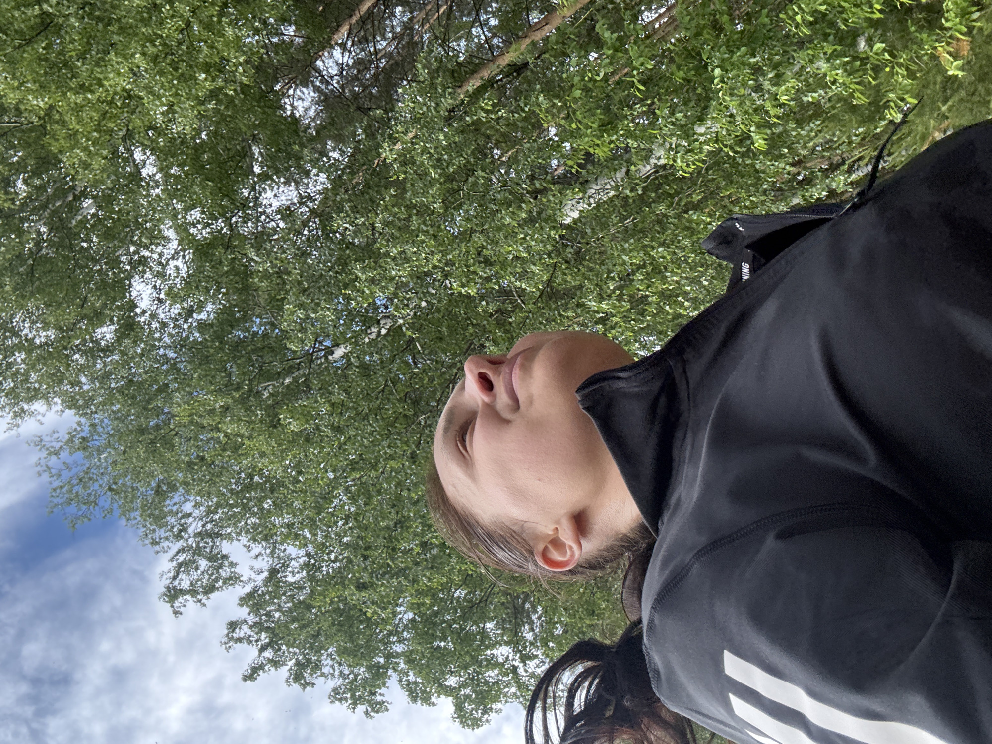This image, originating from Chaikovsky, Russia, presents a low-angle view of an adult female looking upwards in a wooded outdoor environment. The individual is positioned in the lower portion of the frame, with her head and torso visible. She has dark hair, which appears pulled back, and her face is directed upwards and slightly to her right, with a soft, upward gaze. She is wearing a black, high-collared athletic jacket or top featuring distinctive white horizontal stripes on the upper left arm (viewer's right) and a small white label with the partial word "NING" near the collar on the right side of her chest (viewer's left).
The background is dominated by a dense canopy of vibrant green leaves from numerous trees, primarily identifiable as deciduous species like birches due to visible white bark on some trunks. Interspersed among the foliage are clear patches of light blue sky with scattered white, wispy clouds. The perspective is from beneath the tree line, emphasizing the height and density of the forest. The scene is illuminated by daylight, indicative of either late spring or summer. No other people or specific objects are discernible beyond the individual and the natural environment.