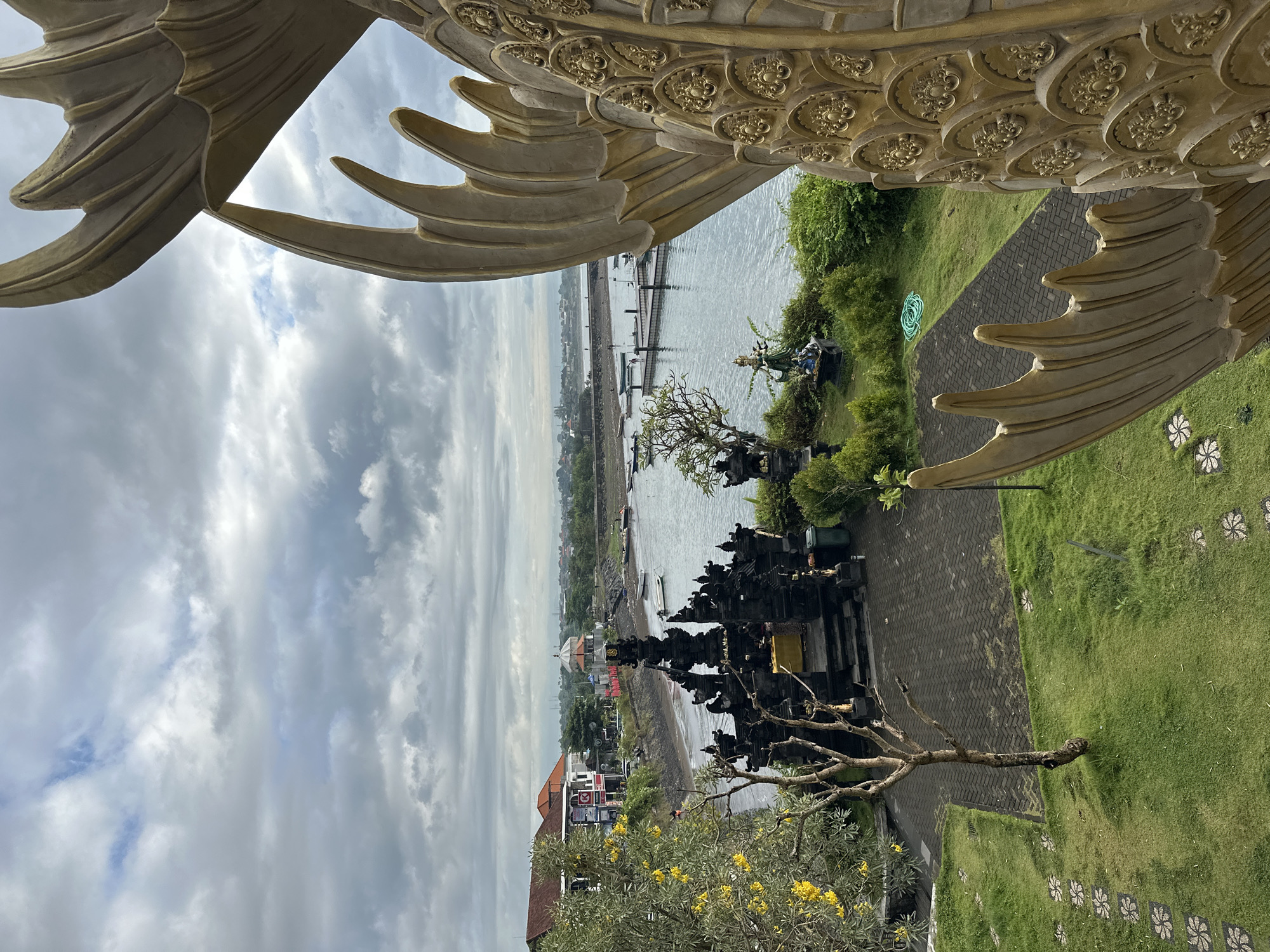 The image presents an elevated view of a scene in Denpasar, Indonesia, capturing a landscape that combines cultural elements with natural surroundings. In the foreground, ornate golden architectural details, possibly belonging to a temple or a similarly designed structure, frame the view. Below, a meticulously maintained green lawn dotted with floral patterns in the grass leads to a stone-paved path. Along the path, a complex of dark, traditional Balinese structures, likely part of a temple complex, stands prominently. In the midground, a body of water stretches towards the horizon, with a pier extending into it. Further in the distance, a coastal city skyline blends into the cloudy sky. Buildings with red-tiled roofs are visible among the trees, indicating urban development in the area. The sky is overcast with varied cloud formations, suggesting a dynamic weather condition.
