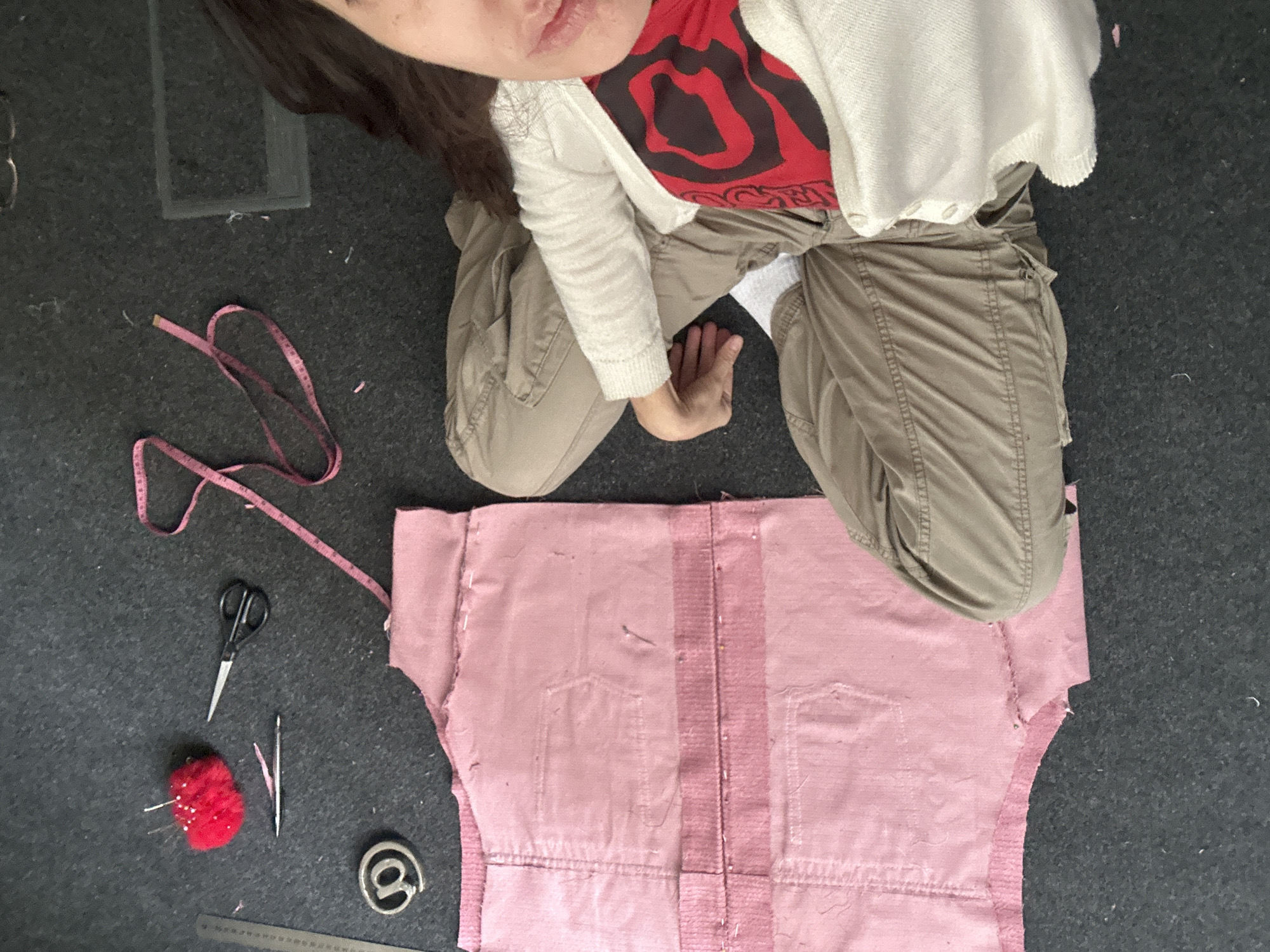 A woman is sitting on the floor, legs crossed, with a partially sewn pink garment in front of her. She is wearing khaki pants, a red shirt with black patterns, and a cream-colored cardigan. To the left of the garment are sewing tools: a pink measuring tape, scissors, a pin cushion with pins, and a metal object resembling an "@" symbol. A ruler is partially visible along the bottom edge. The floor is dark gray. There is no evidence to determine the city or country.