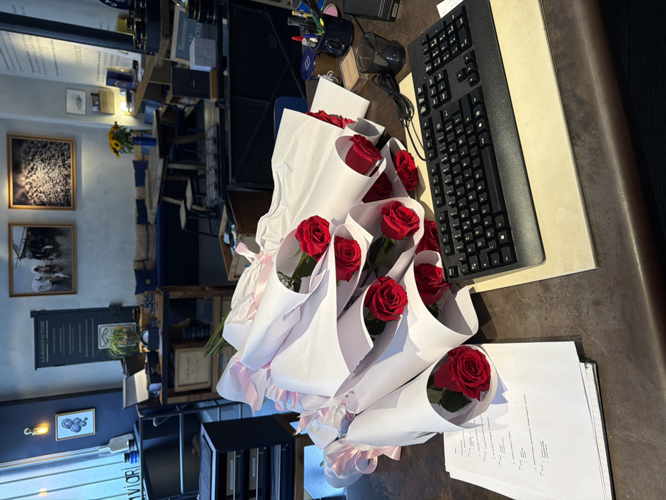 The image depicts the interior of a commercial establishment, identifiable as "La Manufacture" through prominent French signage, located in Paris, France. The foreground is dominated by a dark brown countertop piled with at least twelve individually wrapped red roses. Each rose is presented in white paper, secured with a light pink ribbon. To the right of these roses, a black computer keyboard rests on a light beige mat, accompanied by a black pen holder containing various writing implements and a dark glass. A stack of white documents with printed text lies to the left of the rose arrangements.
The midground reveals a well-appointed showroom or lounge area. On the left, a multi-drawer cabinet is situated near a window with vertical blinds, alongside a wall-mounted light fixture and a partially visible sign with "aviari." The central space features a wooden table with two wooden stools, backed by a long dark blue upholstered bench. Above the bench, two framed artworks hang on a light blue-grey wall: one portraying figures and horses, the other an abstract or textured black-and-white composition. Further right, various retail display items are visible on shelves and tables, including a vase of yellow sunflowers. French text on a wall panel reads "LA MANUFACTURE," and another partially obscured sign says "BIENVENUE À LA MANUFACTURE" (Welcome to the Manufacture). No individuals are present in the scene.