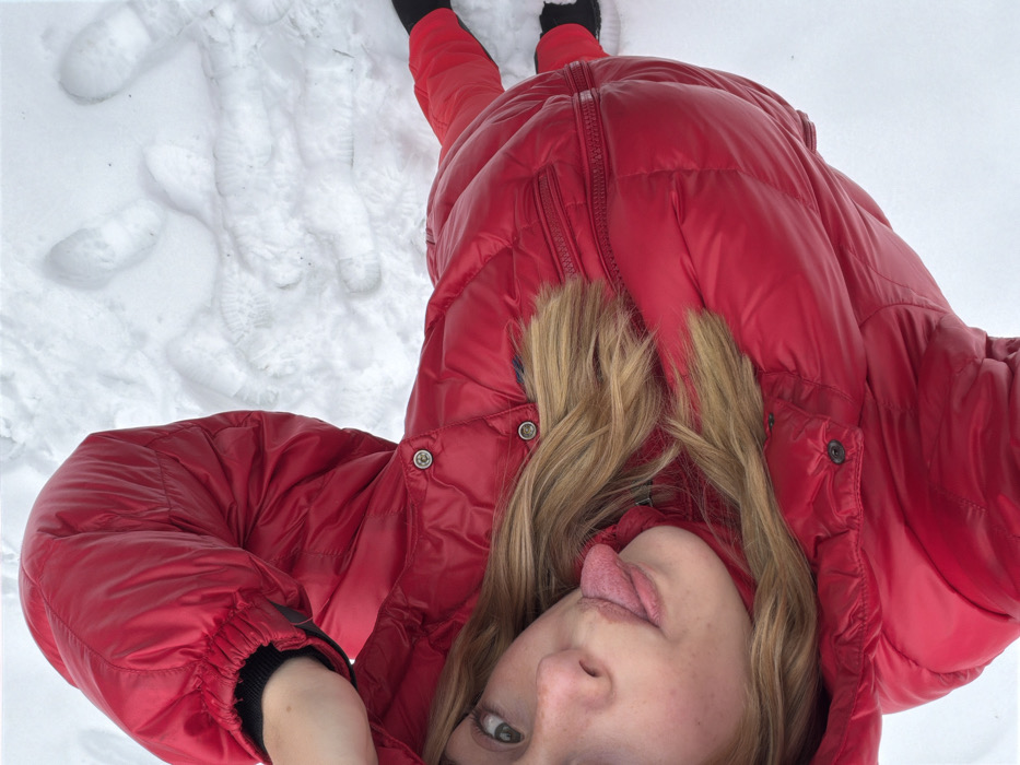 A single individual is depicted lying supine on a snow-covered surface. The subject is wearing a padded, glossy red winter jacket featuring a hood with visible dark fur trim around the neck, and matching red ski pants. Black wrist cuffs are visible on the jacket sleeves, along with black footwear. The individual's head is tilted, revealing light brown hair and a direct gaze towards the camera. The tongue is extended from the mouth. The subject's left arm, clad in a black glove, is raised and positioned near the top of the head. Multiple footprints are discernible in the surrounding snow. The scene is located in La Clusaz, France.