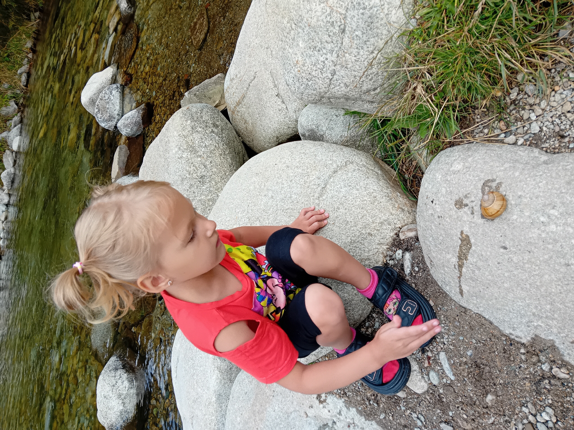 A young girl with blonde hair tied in a ponytail, wearing a red t-shirt with a colorful graphic and black shorts, is crouching on a large boulder next to a clear stream. She is wearing black sandals with pink socks. Her left hand is reaching towards a snail on a rock in front of her. The stream flows over a bed of pebbles and larger stones. Several large, smooth boulders are scattered around the stream bank. Green grass grows in patches near the boulders. The scene appears to be an outdoor natural setting, likely in the vicinity of Zuberec, Slovakia, given the characteristic landscape of the region.