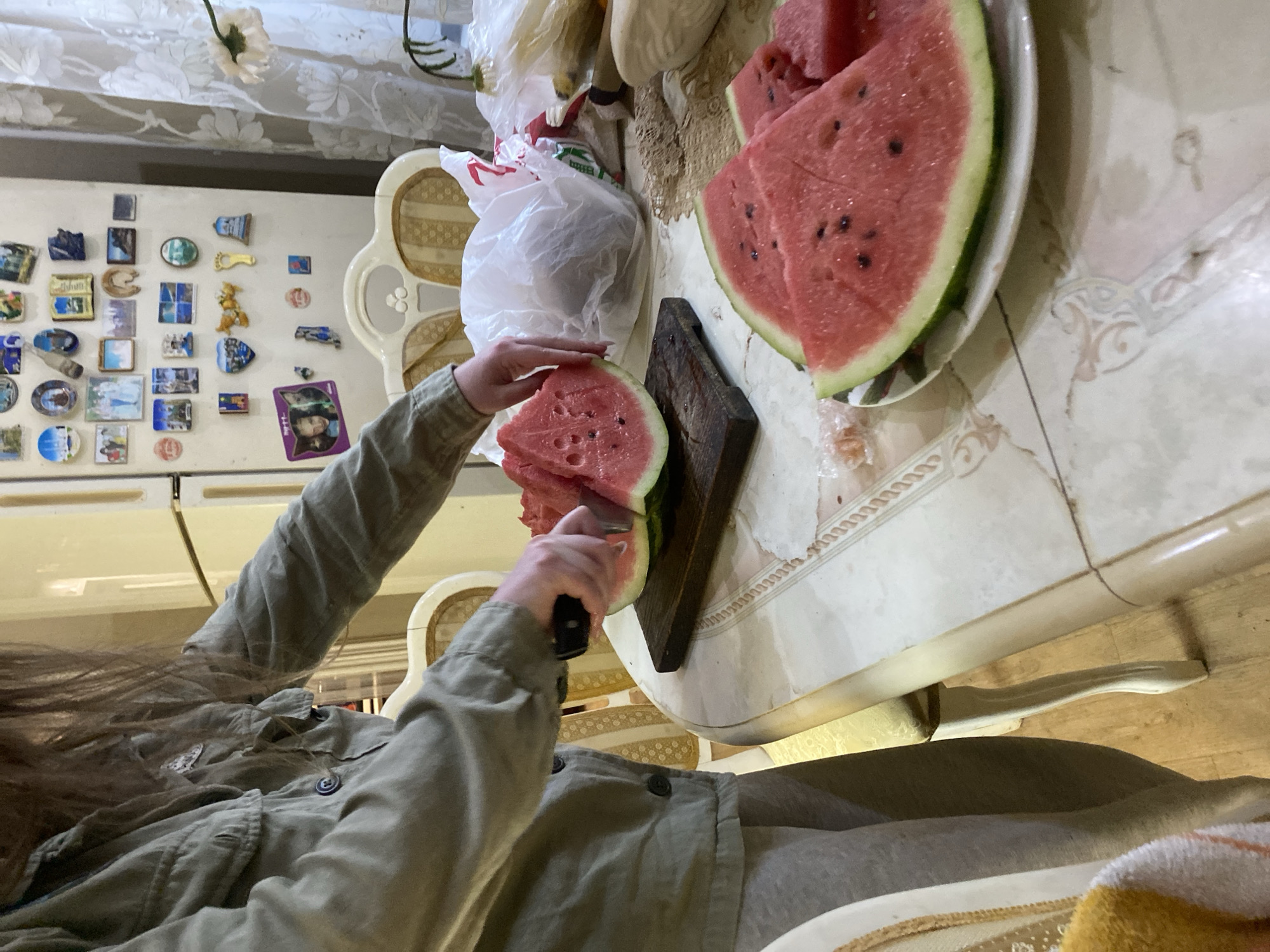 In an indoor kitchen, a person with long brown hair, wearing a grey jacket and grey sweatpants, is cutting a watermelon. The watermelon is on a dark wooden cutting board. Several slices of watermelon are already on a white plate in the foreground. The kitchen features a beige and white refrigerator covered in magnets, a table with a patterned tablecloth, and chairs. Plastic bags and other items are visible on the table. The overall scene suggests a domestic setting, possibly in the city of Volzhsk, Russia, given the presence of what appear to be Russian refrigerator magnets.