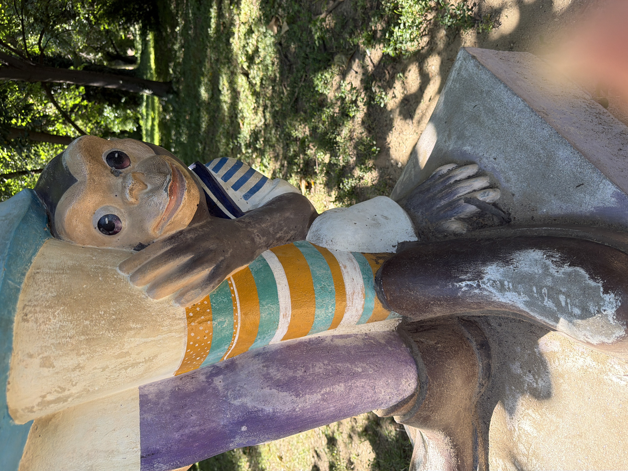 This is a photograph of a statue of a monkey peeking out from behind a larger, possibly human-like figure. The monkey statue has a light brown face with dark, large eyes and a small mouth. It is wearing a blue and white striped collar. The larger figure's leg is visible, painted with stripes of purple, yellow, green, and white. The statue is outdoors, with a grassy area and trees in the background, suggesting a park or garden setting. The photograph appears to be taken in the city of Pazardzhik, Bulgaria, specifically in the area of the Monument of Freedom.