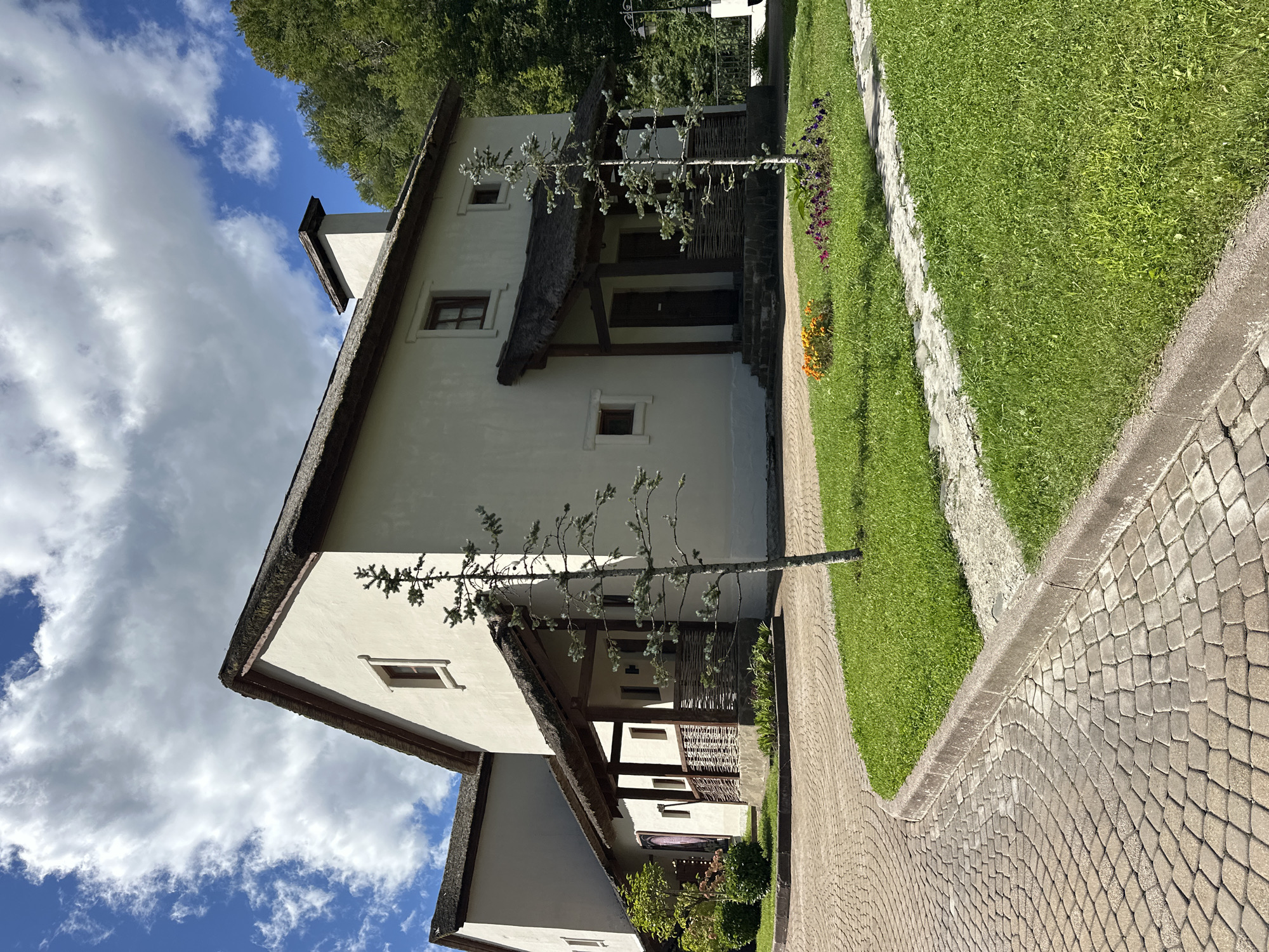The image depicts a white, two-story building with a thatched roof, characteristic of a resort or private residence. The architecture suggests a rustic or traditional style. The building is situated in a lush green environment with mature trees visible in the background. A paved pathway, made of interlocking stones, curves around the front of the building, bordered by a well-maintained lawn with flowerbeds. A thin, young coniferous tree stands prominently in front of the house. The sky is partly cloudy with a mix of blue sky and white clouds. The scene appears tranquil and well-kept, likely part of a larger complex given the architectural style and landscaping. The location is identified as Estosadok, Russia.