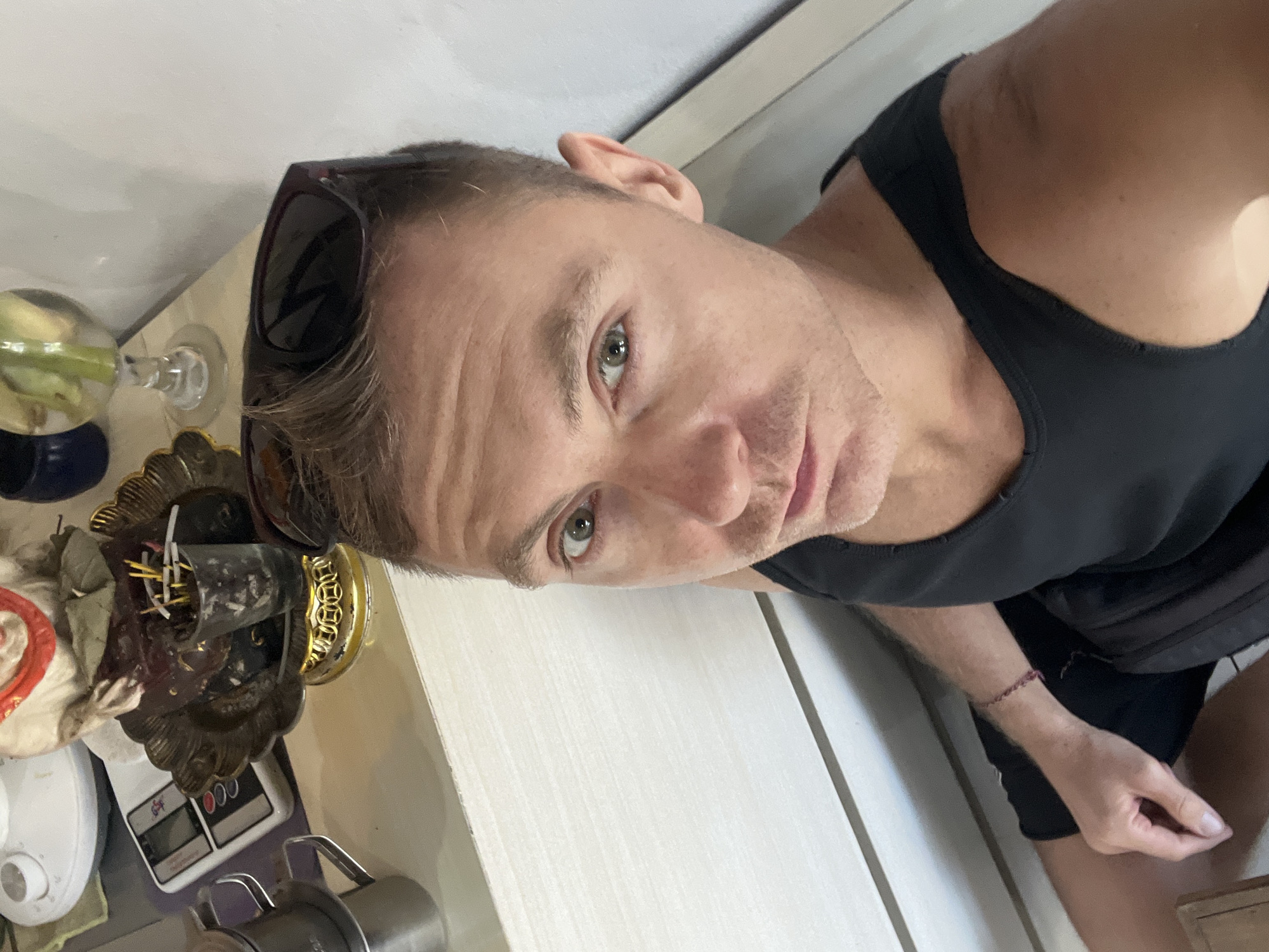 A close-up selfie shows a person in a black tank top, seated and looking directly at the camera. The person has short, light brown hair styled upwards and is wearing sunglasses on their head. Their left hand rests on their leg, and a thin, dark bracelet is visible on their wrist. Behind the person, a white counter or shelf is visible, with several objects including a digital scale, a golden ornate dish with incense sticks and other items, a small metal pitcher, and a glass with a drink. The background appears to be a white wall. The scene is likely indoors, possibly in Kuta Selatan, Indonesia, given the context of the objects and the tropical lighting.