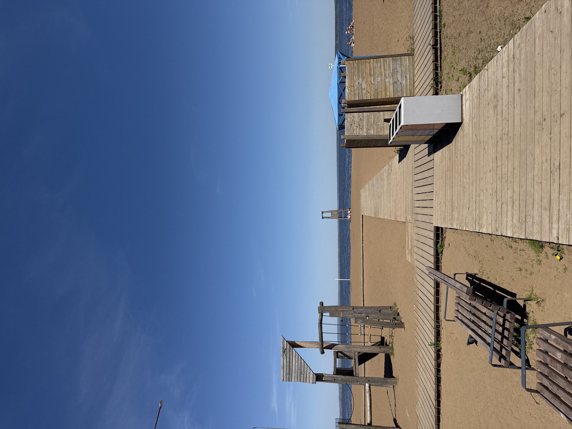 The image depicts a beach scene in Chaikovsky, Russia, characterized by a sandy shoreline, a blue sky, and several wooden structures. On the right, a wooden boardwalk runs alongside a grassy area, leading towards the beach. A wooden bench is located near the boardwalk. Further towards the beach, there is a wooden changing stall with a blue roof. Another wooden structure, possibly a lifeguard station or part of a playground, is visible on the left. The presence of people on the beach, albeit in the distance, indicates recreational use. The scene suggests a public beach area for leisure activities and relaxation.