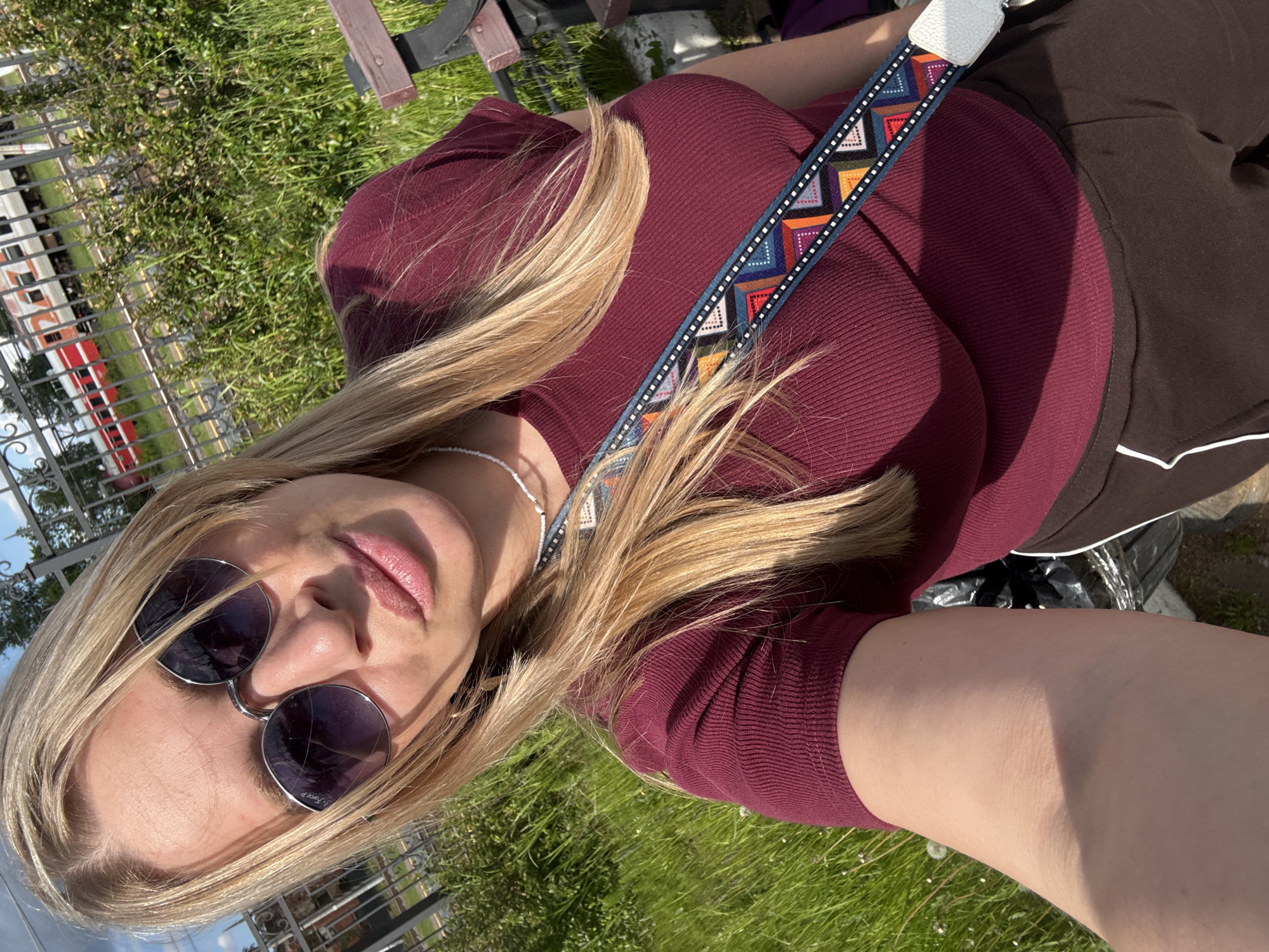 A bright, angled outdoor selfie features a woman with long, sun-kissed blonde hair, fair skin, and plump pink lips, wearing round sunglasses. The text "P-Force P" is visible on the left lens of her sunglasses. She is dressed in a close-fitting, ribbed maroon or burgundy top and dark brown pants or a skirt with white trim details. A prominent, multicolored patterned strap, decorated with geometric shapes in hues of blue, orange, red, and white, crosses her torso. A delicate white beaded necklace is also visible around her neck.
The setting is outdoors, characterized by vibrant green grass and partial sunlight, indicating a sunny day. In the background, behind a decorative black metal fence, a train with red and white carriages is partially visible, suggesting the photo was taken near a railway line or station. A wooden bench is also present in the upper left corner of the frame. The overall scene depicts a casual moment outdoors on a clear day, within the context of Malaya Vishera, Russia.