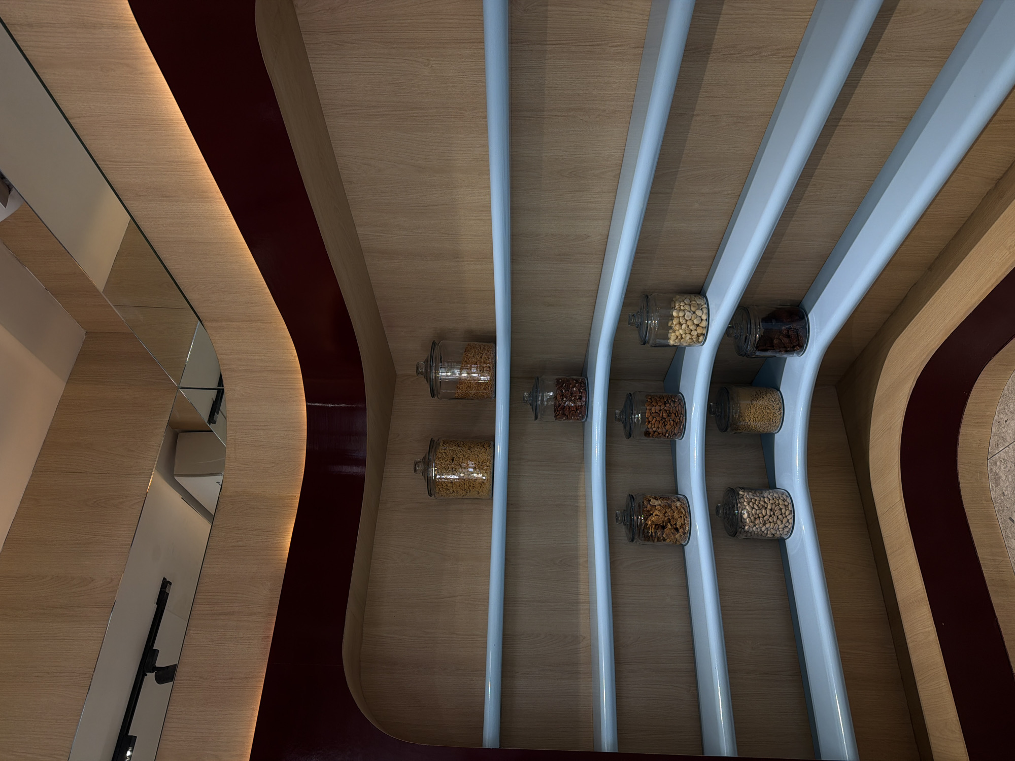 The image captures an abstract interior design element. Wooden curved walls form a pattern with blue vertical stripes, creating compartments where glass jars filled with different kinds of nuts and dried fruits are displayed. A mirrored section reflecting an interior space is visible on the left. The location appears to be in Mexico City, Mexico, based on the architecture and design style. No people are visible in the scene.