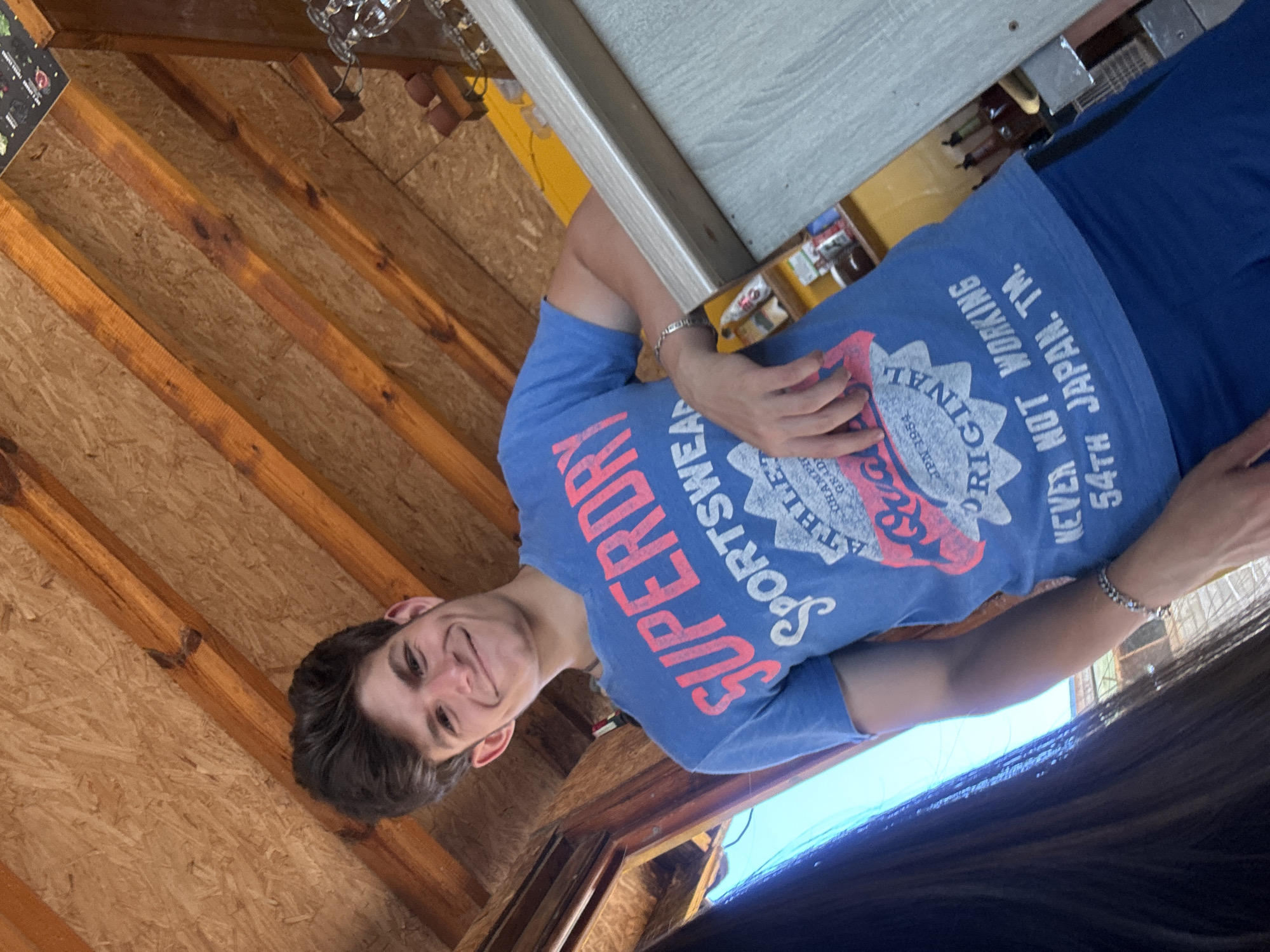 The image captures a young man with dark, tousled hair and a broad smile, posing indoors. He is wearing a blue t-shirt with the "SUPERDRY" logo prominently displayed in red and white lettering, along with text that reads "SPORTSEWER ATHLETIC GRADED ORIGINAL JPN 1954 NEVER NOT WORKING 54TH JAPAN. TM." He is also wearing blue shorts. The man is leaning against a counter with a light-colored surface. Behind him, the ceiling is constructed of wooden beams and plywood. To the left, a person's dark hair is visible, partially obscuring the view of the man's left side. The background shows shelves with bottles and other items, suggesting a bar or shop environment. The overall lighting is bright, indicating it's daytime. The style of the ceiling suggests a rustic or beach-themed establishment, which is consistent with the atmosphere often found in coastal towns like Sozopol, Bulgaria, known for its historic architecture and seaside attractions.