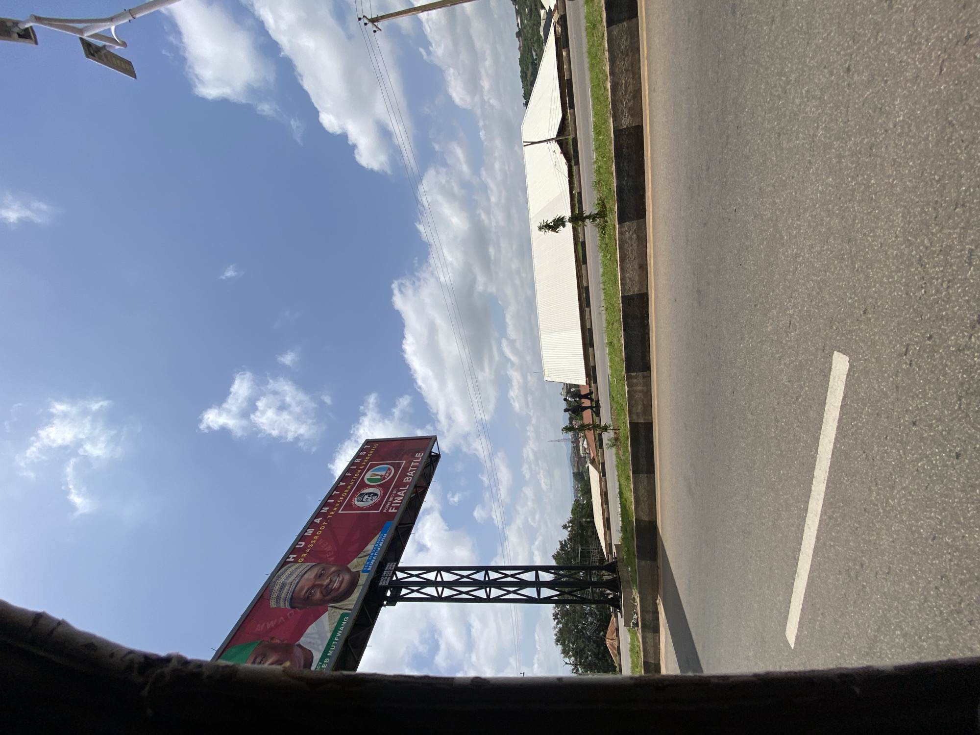 This image captures a street scene in Jos, Nigeria. A large billboard dominates the upper left portion of the frame, displaying images of two men, one wearing a green cap and the other a patterned cap and a green shirt. Text on the billboard reads "HUMANITY FIRST GRASSROOT TRANSFORMATION & PROGRESS" and "FINAL BATTLE". Below the billboard, a metal support structure is visible.
In the background, a long, white-roofed building stretches across the landscape. A power pole with a solar panel is positioned on the right side of the image, and power lines are strung across the sky. The foreground shows a paved road with a white line marking a lane. A concrete barrier with black and gray bands separates the road from a grassy area. Several individuals are walking in the middle ground, near the white-roofed building. The sky is blue with scattered white clouds. The overall scene suggests an outdoor urban or semi-urban environment in Jos, Nigeria.