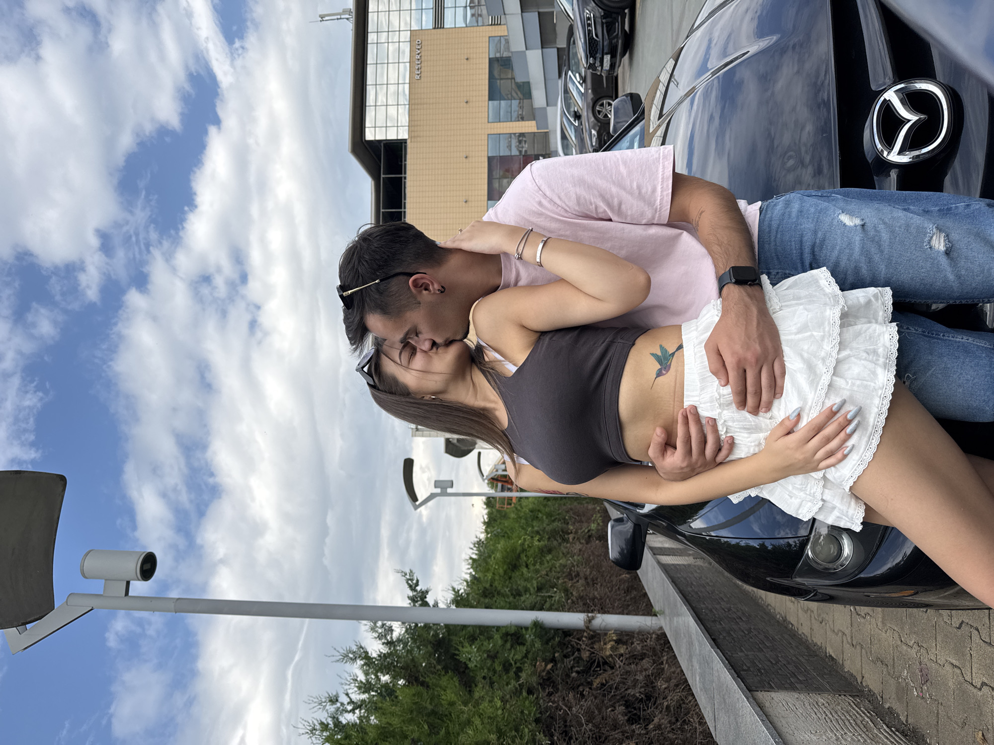 The image depicts an outdoor scene in Sofia, Bulgaria, featuring a couple embracing and kissing in front of a black Mazda car. The location is identified by the "RESERVED" store visible in the background and a Bulgarian license plate (CA prefix for Sofia) on a Kia SUV parked further back.
The female, positioned on the left, has long brown hair, dark sunglasses perched on her head, and is wearing a dark grey crop top, a white ruffled mini-skirt, and displays a hummingbird tattoo on her left lower abdomen. Her left arm is around the male's neck, and her right hand rests on his chest. Her fingernails are long and painted light grey.
The male, on the right, has dark hair with sunglasses on his head, and is wearing a light pink short-sleeved t-shirt, ripped blue jeans, and a black smartwatch on his left wrist. He has earrings in his left ear. His left arm is around the female's waist, with his right hand on her hip/thigh area.
The couple is in a close embrace, engaged in a kiss.
In the foreground, a black Mazda car's hood and front grille are prominent. Behind the couple, a paved parking area contains several other parked vehicles, including a black Kia SUV with a visible "CA" Sofia license plate. A modern commercial building with glass windows and a beige facade, bearing the "RESERVED" signage, forms the primary background structure.
To the far left, a dense hedge of green coniferous bushes runs along a low stone wall and a tiled pedestrian walkway. Several grey streetlights are visible, including a prominent one in the upper left of the frame. The sky is partly cloudy with patches of blue.