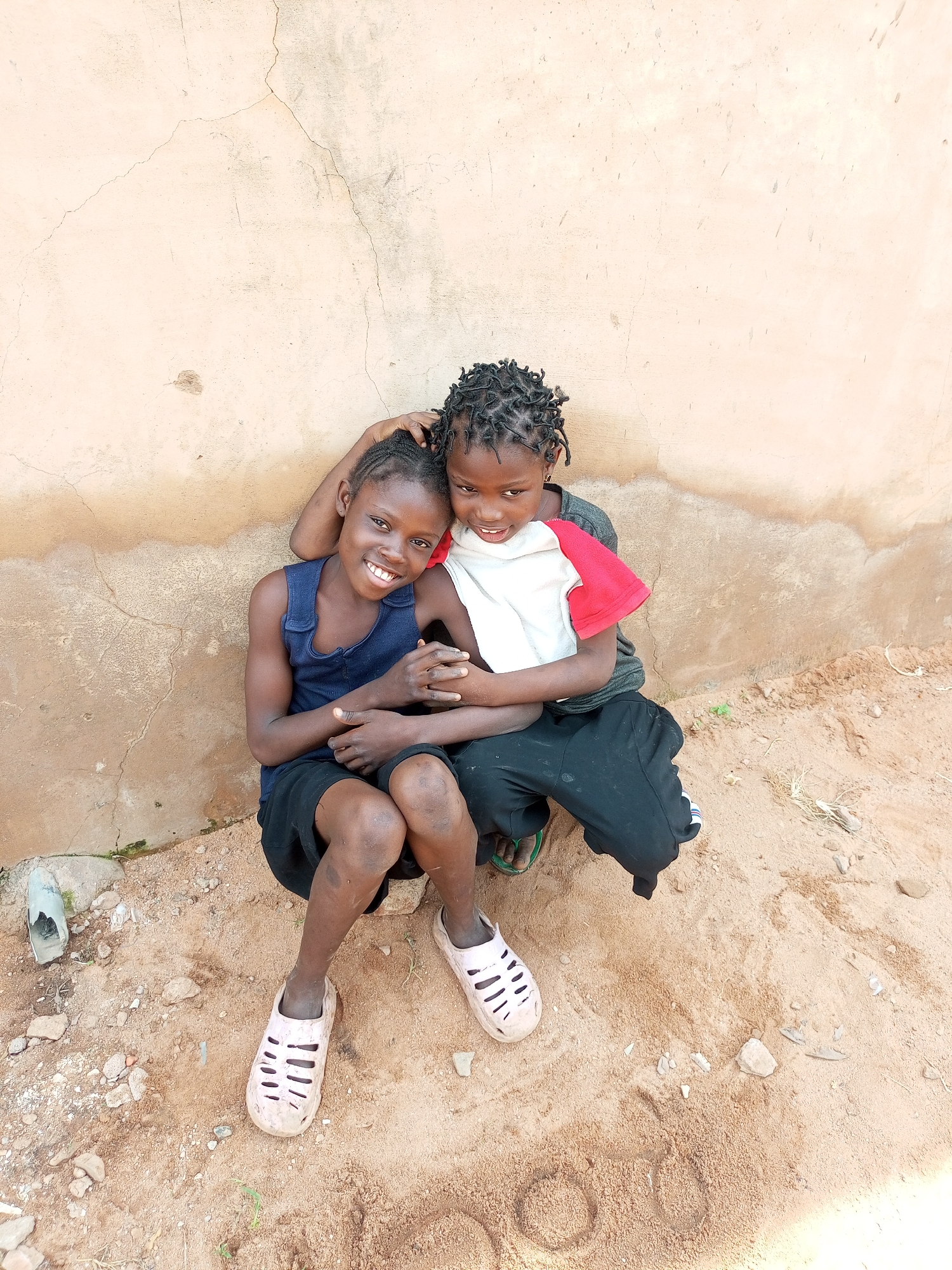 Two children are seated on sandy ground against a textured, light-colored wall in Tempure, Nigeria. The child on the left, wearing a dark blue tank top and black shorts, is looking at the camera with a broad smile. The child on the right, wearing a gray t-shirt with red sleeves, has their arm around the other child and is also smiling. Both children have their hair in braids. The ground beneath them is sandy and contains small stones and some dry vegetation. The wall behind them shows signs of weathering and has some cracks.