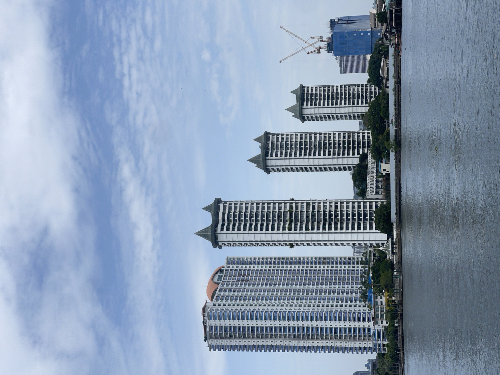 The image depicts a waterfront view of the Chao Phraya River in Bangkok, Thailand, with a skyline dominated by several high-rise buildings. On the left, a very tall, white apartment building with blue balconies stands prominently, topped with a curved, orange roof. Next to it, to its right, are two similar, slightly shorter, white high-rise buildings, each featuring a distinctive green, pointed roof structure reminiscent of a castle turret. Further to the right, another, even shorter building with the same turret-like roof design is visible. In the far right of the frame, a construction site is evident, with two tall cranes extending above a blue-covered building under construction. The foreground is occupied by the calm, greyish-blue water of the river. Along the riverbank, a line of trees and various smaller structures, including a white pavilion, are visible. The sky above is partly cloudy, with patches of blue sky interspersed with white clouds. No people are clearly discernible in the image. The overall impression is one of urban development along a major river.