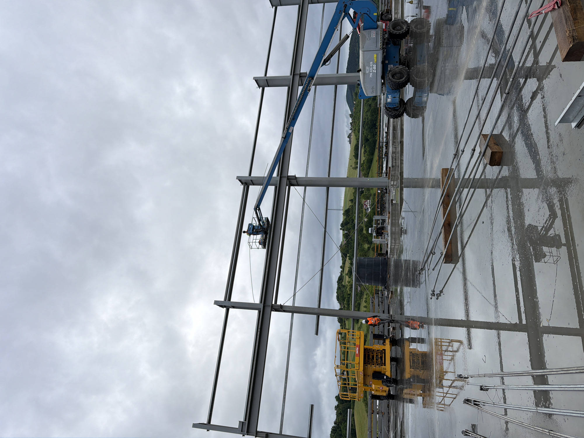 The image depicts a construction site in Janowice, Poland, under a cloudy sky. The foreground shows a wet, reflective concrete surface with various construction materials, including metal rods and wooden blocks. In the midground, a yellow scissor lift and a blue boom lift are positioned near a large steel structure that forms the framework of a building. A worker in an orange vest and hard hat stands near the scissor lift, while another worker is visible in the basket of the boom lift. In the background, rolling hills covered in green trees and dotted with houses suggest a rural or semi-rural setting. The overall scene conveys an active construction phase with heavy machinery and workers engaged in building operations.