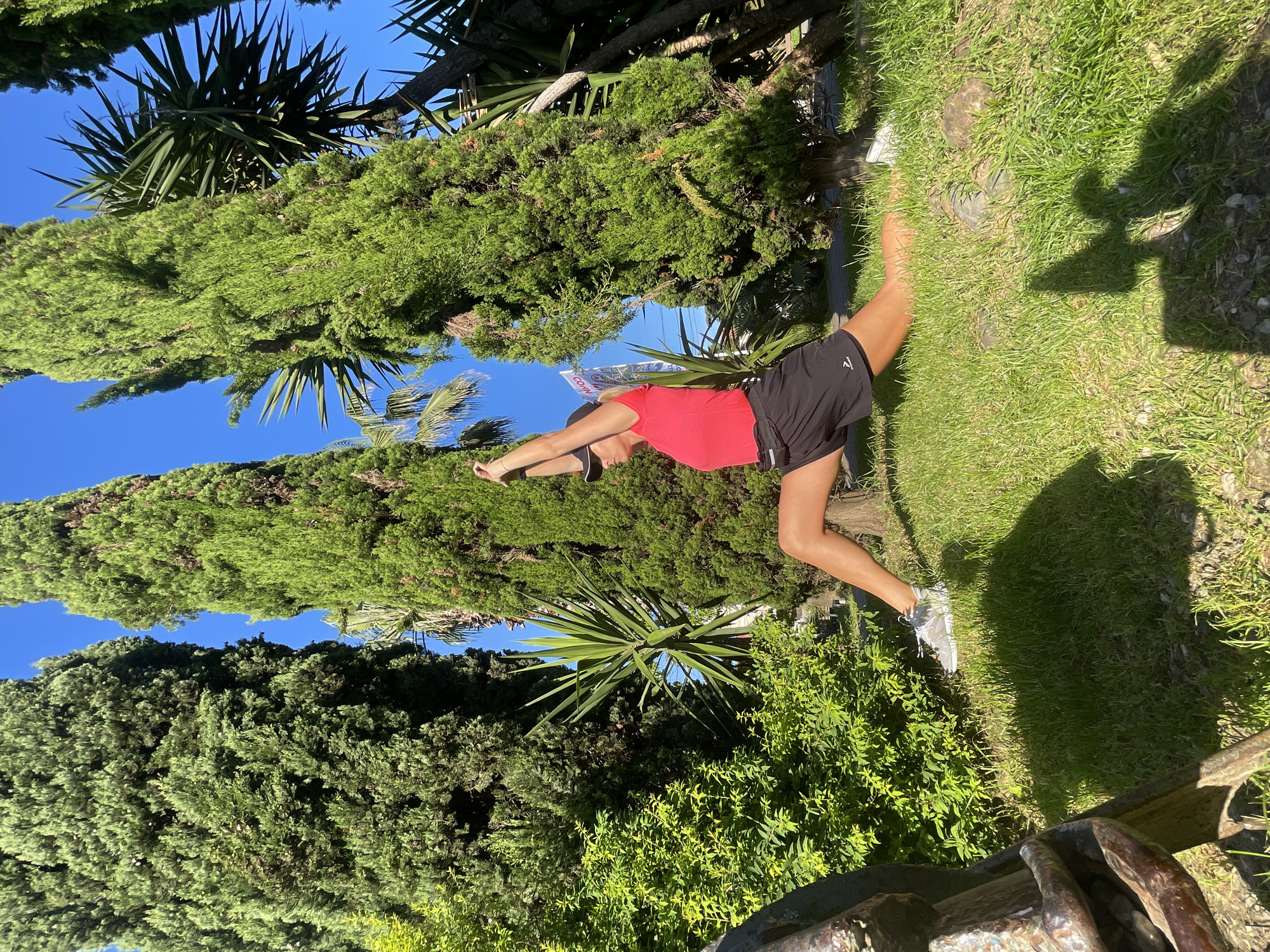 This outdoor image, originally rotated 90 degrees clockwise, captures a woman stretching or exercising on a bright, sunny day in Sochi, Russia. The woman, positioned centrally, is dressed in a vibrant red short-sleeved top, black athletic shorts with a white stylized 'A' or arrow-like logo on the left thigh, a dark baseball cap, and light-colored sneakers. Her arms are raised and extended forward with hands clasped, as she leans slightly, indicative of a warm-up or stretching movement.
The setting is a lush, green environment under a clear, intense blue sky. Tall, slender coniferous trees, possibly cypress or juniper, rise prominently in the background and on either side, their dense foliage providing a rich green backdrop. Among these, spiky-leaved yucca or palm-like plants are also visible. The ground beneath the woman is covered in well-maintained green grass, interspersed with some visible rocks. Strong, elongated shadows, including those cast by the woman and an unseen object or person, stretch across the grass, suggesting it is either mid-morning or late afternoon. A white banner or sign with unclear red and blue text (partially resembling "COVN") is faintly visible behind her head. A dark, metallic-looking railing or structure is present in the lower right foreground.
