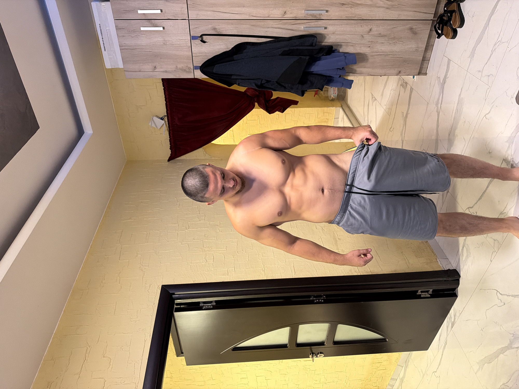 A male individual stands shirtless and barefoot in an indoor room. He is positioned centrally, looking downwards, holding the waistband of his grey athletic shorts slightly pulled down on his right hip, revealing a sliver of skin. He possesses a muscular build, a short buzz cut, and light facial stubble. The room features textured yellow-ochre walls, a white baseboard, and a white ceiling with a modern design including a rectangular recess and a dark grey square panel within it. A dark brown interior door with frosted glass panels is open on the left, leading to another yellow-walled area. On the right, a light wood-grained wardrobe with silver handles has a dark blue jacket, a blue shirt, and a black strap hanging from it. Below the wardrobe, two black platform sandals are on the floor. In the background, a tied-back dark red curtain is visible, with a small white bottle below it. Above the wardrobe, a white box with "PRODUCTION LOT NO. A42805SB230026" printed on it rests. The floor is covered with large, light-colored tiles patterned with dark grey veining. This scene is located in the city of Plovdiv, Bulgaria.