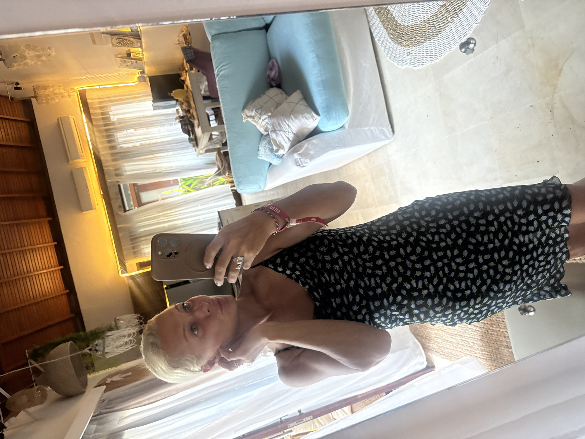A woman with short blonde hair takes a mirror selfie in what appears to be a living room. She is wearing a black slip dress with a small floral pattern. She is holding a smartphone in her right hand and touching her left ear with her left hand. The room has a light-colored tiled floor and white sheer curtains. In the background, there is a light blue couch with several pillows, a dining table with chairs, and a wooden ceiling. There are also hanging lamps and air conditioning units visible. The lighting suggests it is daytime.