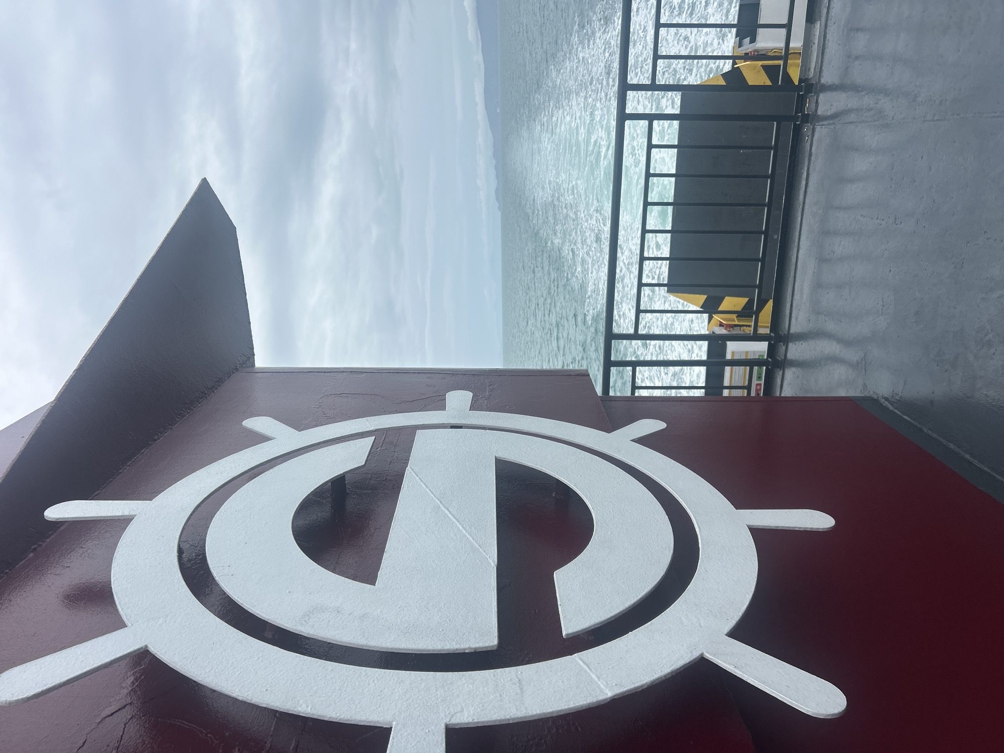 The image shows a close-up of a white stylized "S" logo on a dark red surface, which appears to be part of a ship or ferry. The logo resembles a ship's wheel. In the background, a body of water with a wake is visible under a cloudy sky. A black metal railing runs along the edge of the deck. The overall scene suggests being on a vessel at sea. The image does not provide any information about the specific location in the city of Unknown, Thailand.