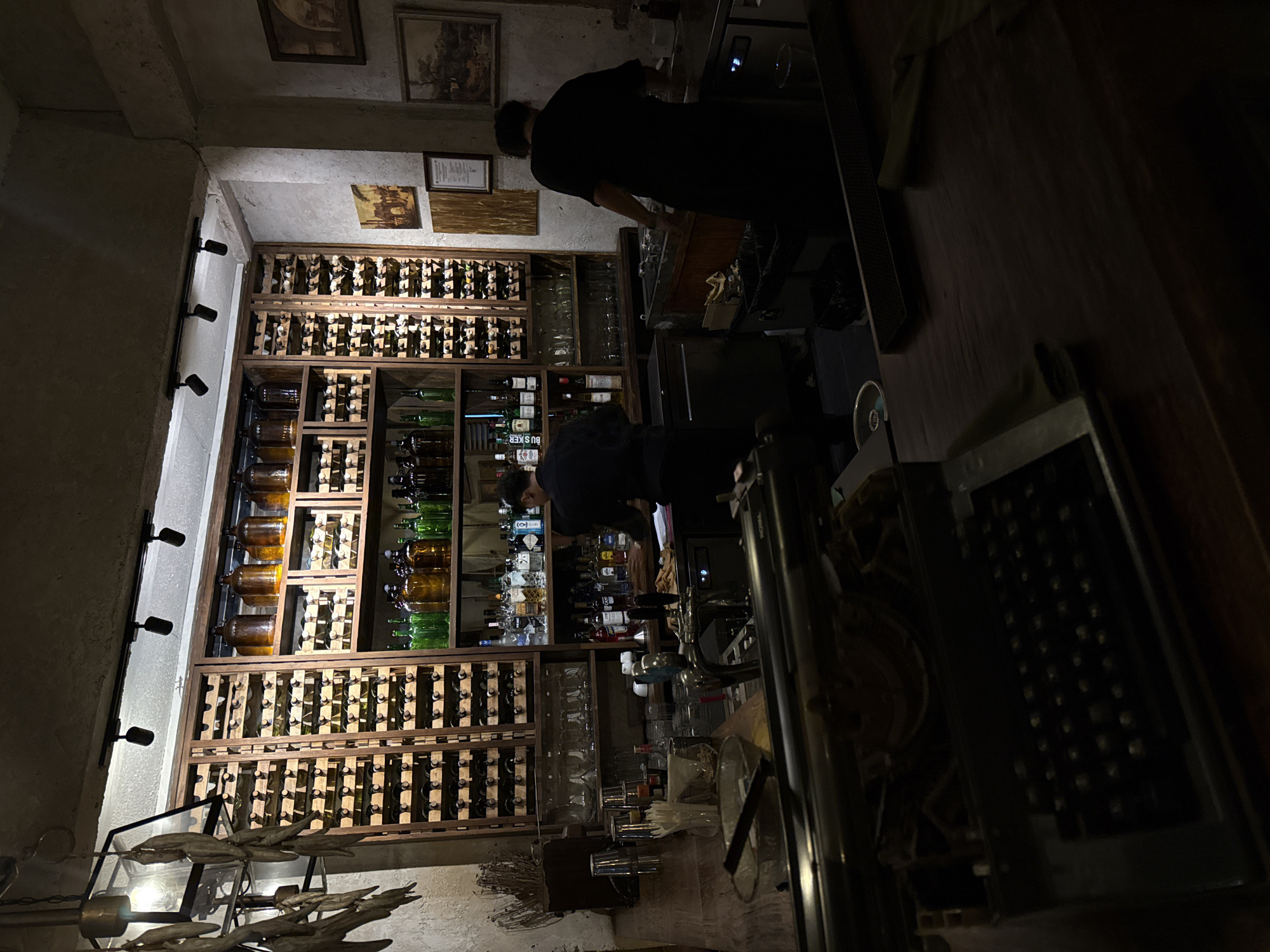 The image depicts the interior of a dimly lit establishment, possibly a bar or restaurant, located in an unknown city. Two individuals, presumably bartenders, are seen working behind the bar. One is standing and appears to be preparing drinks or tending to tasks at the counter, while the other is bending over, possibly reaching for something or attending to equipment.
The background features a large wall-mounted shelving unit filled with numerous bottles of various alcoholic beverages, predominantly wine. The bottles are arranged neatly on wooden racks. Above the shelves, there are framed pictures or artworks hanging on the wall.
In the foreground, there's a partially visible object that appears to be an antique typewriter or similar mechanical device. The lighting is subdued, with only a few spotlights illuminating the shelves of bottles and some sections of the bar area. The overall atmosphere is subdued and intimate.