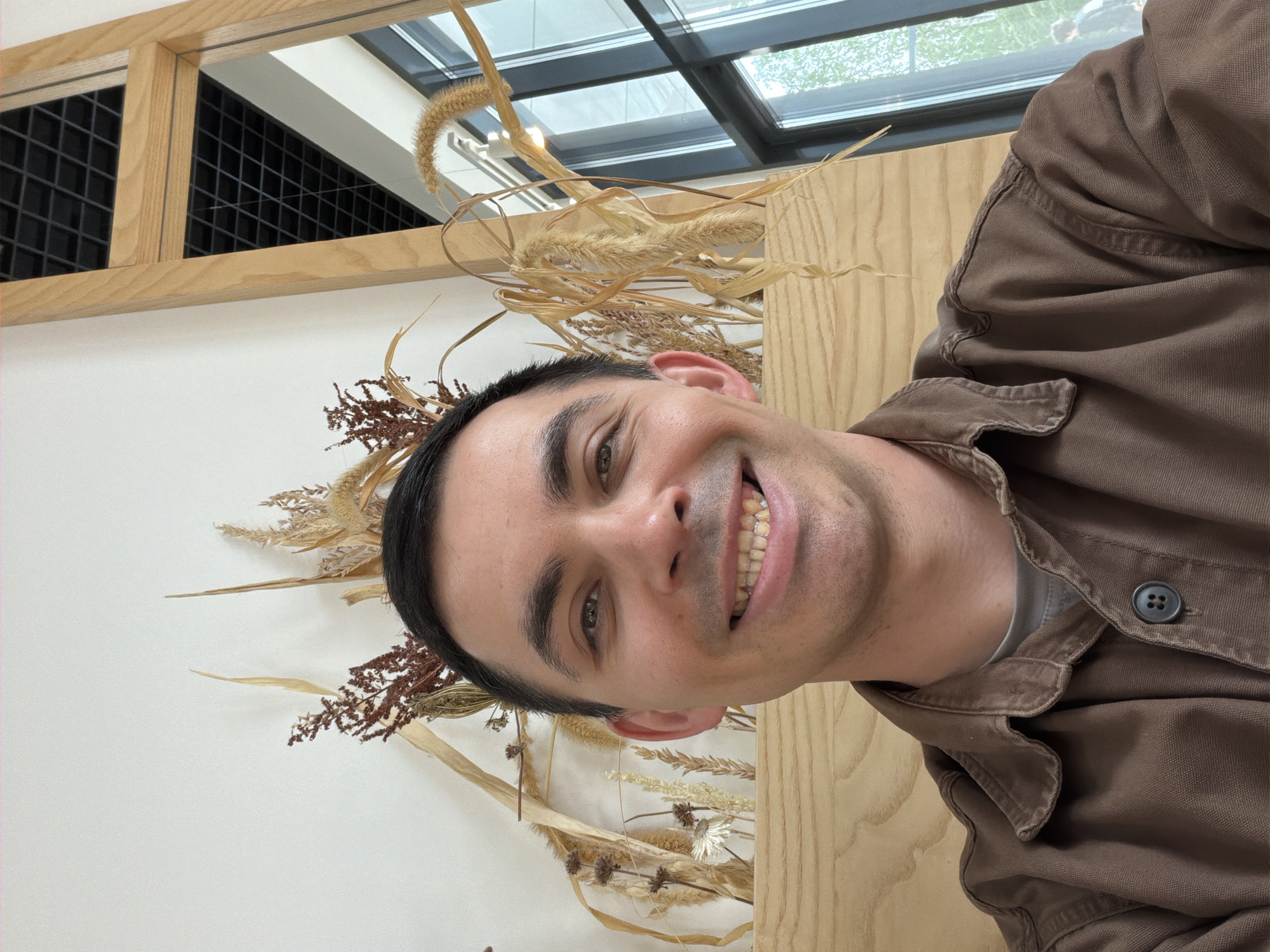 A selfie shows a smiling man with short dark hair and fair skin, wearing a brown button-down shirt. He is partially obscured by light-colored wooden planks and dried floral arrangements that include brown and beige grasses and stems. Above the floral arrangement, a grid-patterned skylight is visible, suggesting an indoor setting, though the exterior is discernible through the skylight. There is no indication of the location being Izhevsk, Russia.
