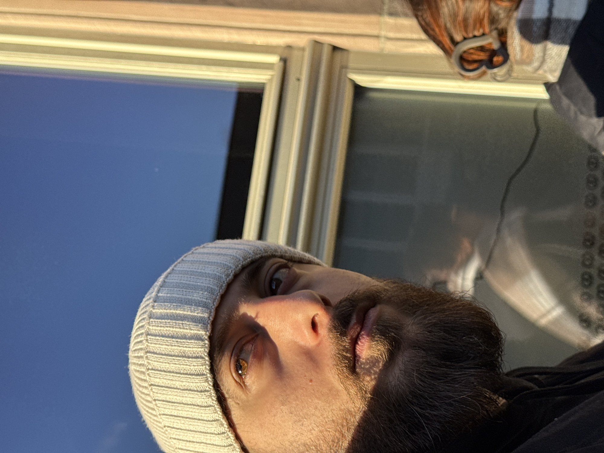 The image captures two individuals outdoors in Strasbourg, France, illuminated by strong, warm sunlight. In the foreground, a male with a full dark beard and a light-colored ribbed beanie is seen from the chest up, looking upwards and to the right. His face is brightly lit, highlighting an amber-brown eye. To his right and partially visible, the back of a second individual's head and shoulder show reddish-brown hair with a heart-shaped hair accessory, and a light-colored plaid garment. The background features a clear blue sky on the upper left and a building segment with a window on the right. The window has light-toned frames and displays reflections, including an indistinct patterned reflection in its lower section.