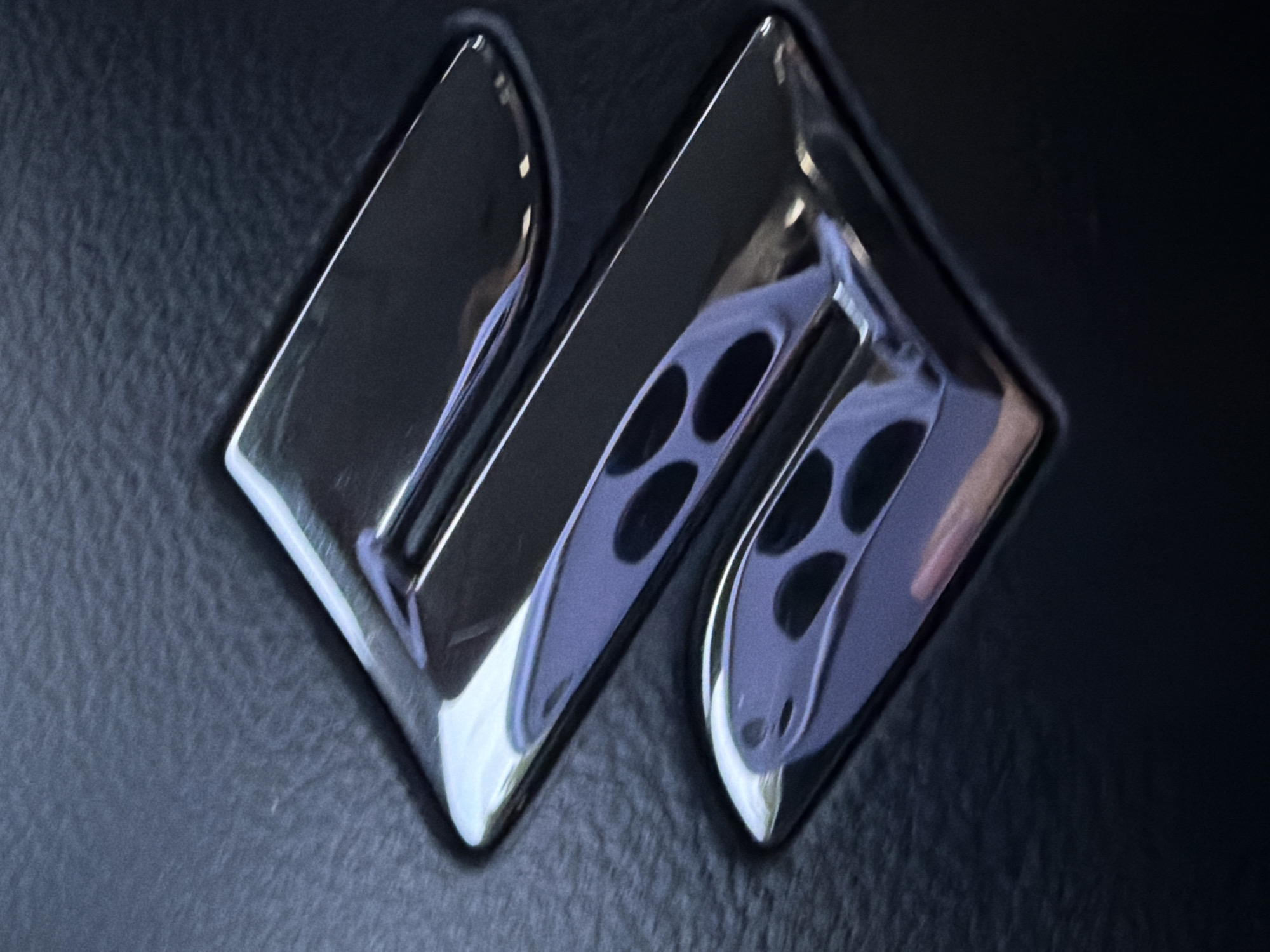 The image displays a close-up of a highly reflective, metallic emblem, prominently positioned against a dark, textured background. The emblem, which strongly resembles the Suzuki automotive logo, is chrome or polished silver in color and features a distinctive stylized "S" or "M" shape, formed by two interlocking angled elements.
The background is a dark, possibly black or very dark grey, textured material, characteristic of automotive interior finishes such as leather or a synthetic equivalent. The emblem's polished surface exhibits clear reflections of its surroundings. Notable reflections include a purple-hued distortion showing a pattern of four dark circular shapes, which could be a car's air vent grille, speaker cover, or similar patterned component. Other reflections show subtle light sources and abstract shapes, possibly hinting at an interior lighting setup or other car cabin elements. A faint, elongated reflection on the right side of the emblem could suggest a hand or finger.
No explicit text is visible on the emblem, but its iconic shape functions as a brand identifier. The scene is static, focusing solely on the object. There are no people visible, and no specific activity is taking place. The overall impression is an interior shot, likely within a vehicle, possibly on a steering wheel, dashboard, or another part of the cabin. The image does not provide specific cues for time of day, weather, or the broader location within Steklyannyi, Russia, beyond the inferred vehicle interior.
