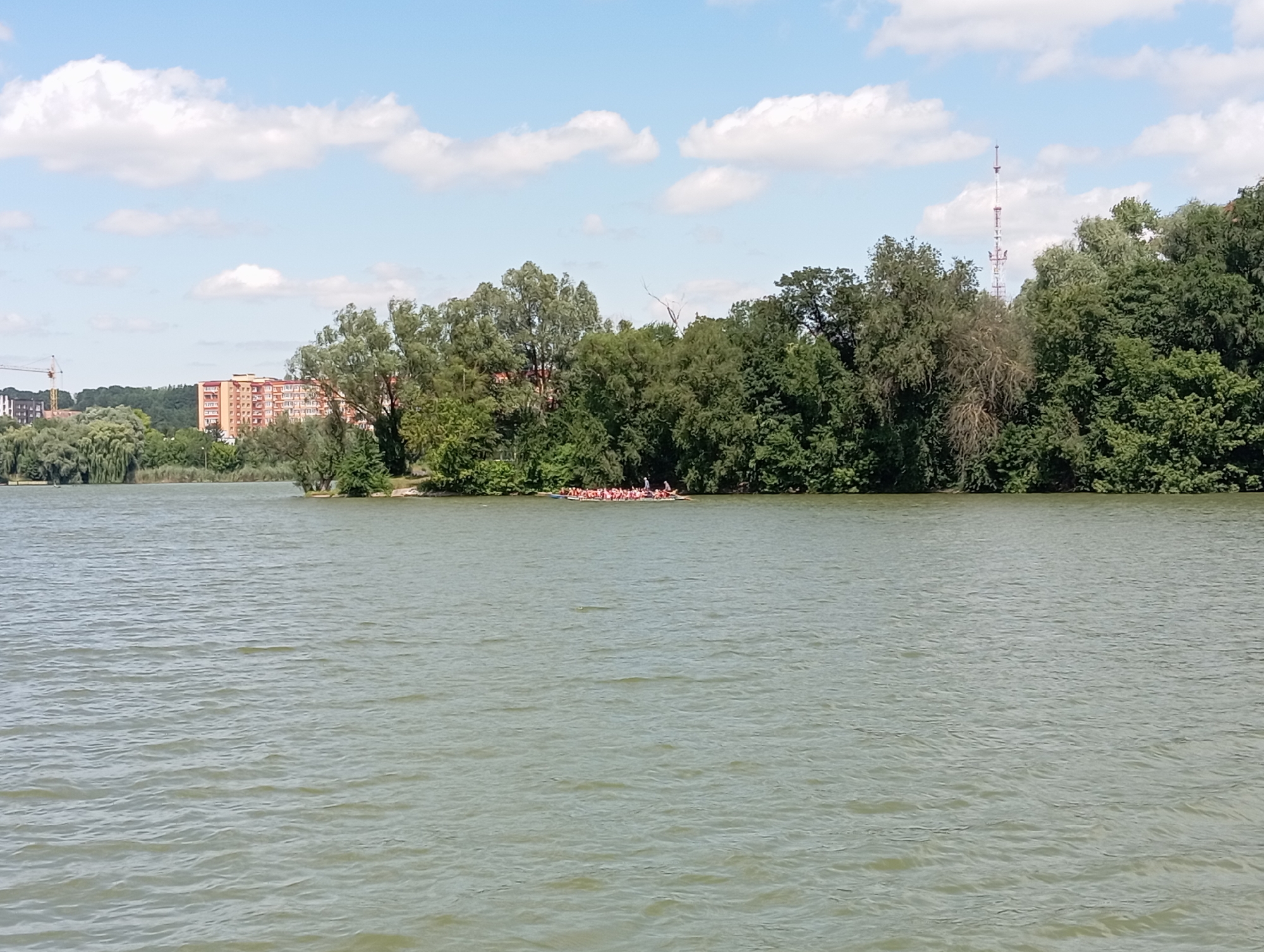 A long, narrow boat filled with people is on a body of water. The boat is long and white with red accents on its sides. There are at least twenty people in the boat, all facing forward. The water is calm with small ripples. In the background, there are trees and buildings. A tower with an antenna is visible in the distance. The sky is blue with white clouds.