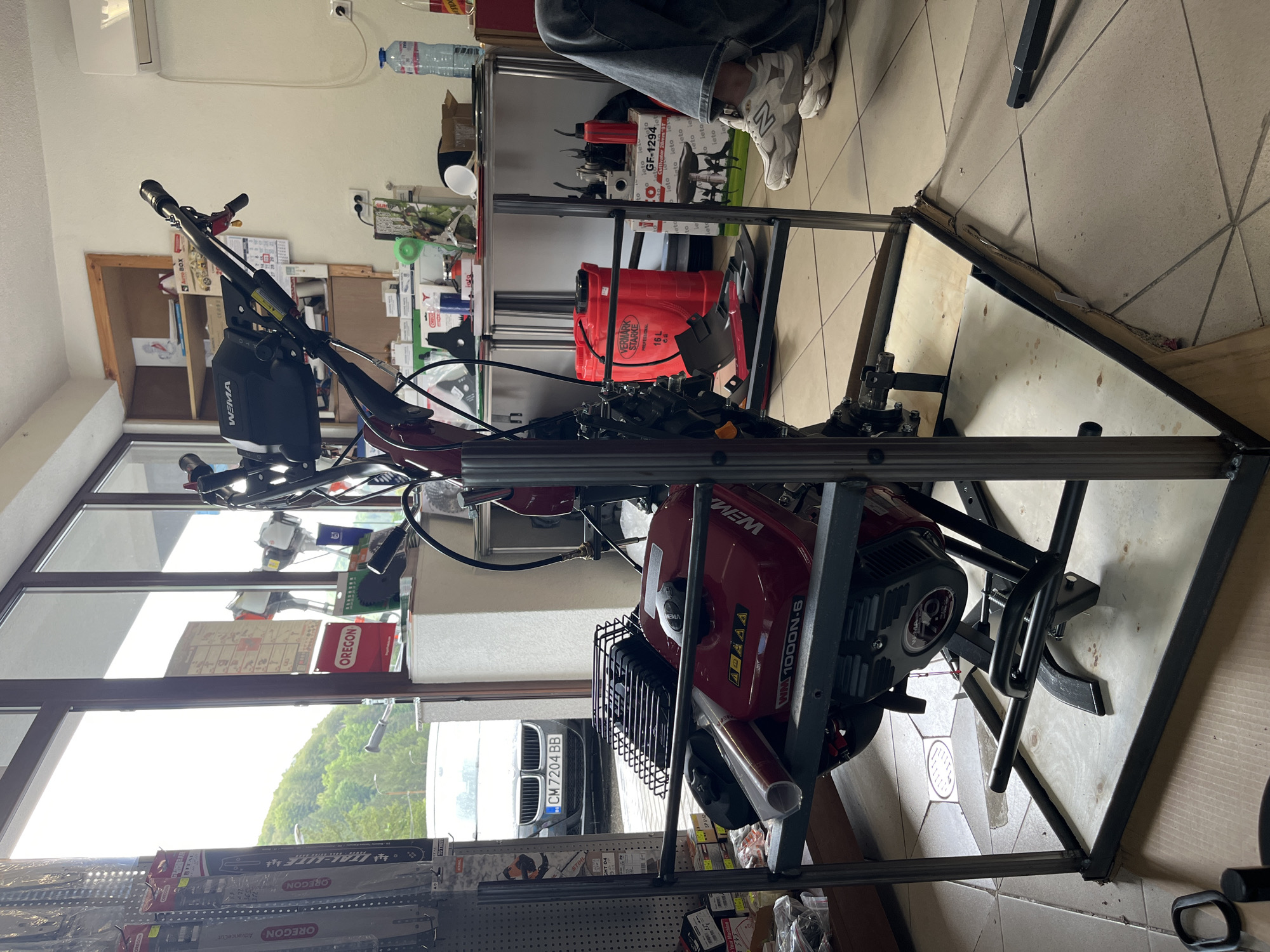 Here is a detailed description of the image:
**Scene:** The image captures a close-up of a red Weima cultivator placed inside a metal frame, seemingly in a workshop or retail environment.
**Objects:**
* The main subject is the red **Weima 1000N-6 cultivator** encased in a custom metal frame.
* **A red VERMARK STARKE 16-L sprayer**
* **A GF-1294 Cultivator 28mm 9T**
* **OREGON chain saw**
* **AUTO BOX**
* Assorted tools, equipment, and retail products are visible on shelves and in the background.
* A white car is parked outside the window.
* The ground is tiled.
**People:**
* A pair of legs wearing jeans and white/grey sneakers are visible in the upper-right corner.
**Environment:** The scene appears to be indoors, likely a workshop, garage, or retail store, as evidenced by the shelving, various tools and equipment, and display items. The presence of windows suggests a well-lit interior.
**Location:**
The image shows the license plate of a car, CM 7204 BB, which suggests the photo was taken in Bulgaria.
**Note:** Due to the limited view and the nature of the image, it's difficult to provide an exhaustive description of every detail, but the provided information covers the key elements present.