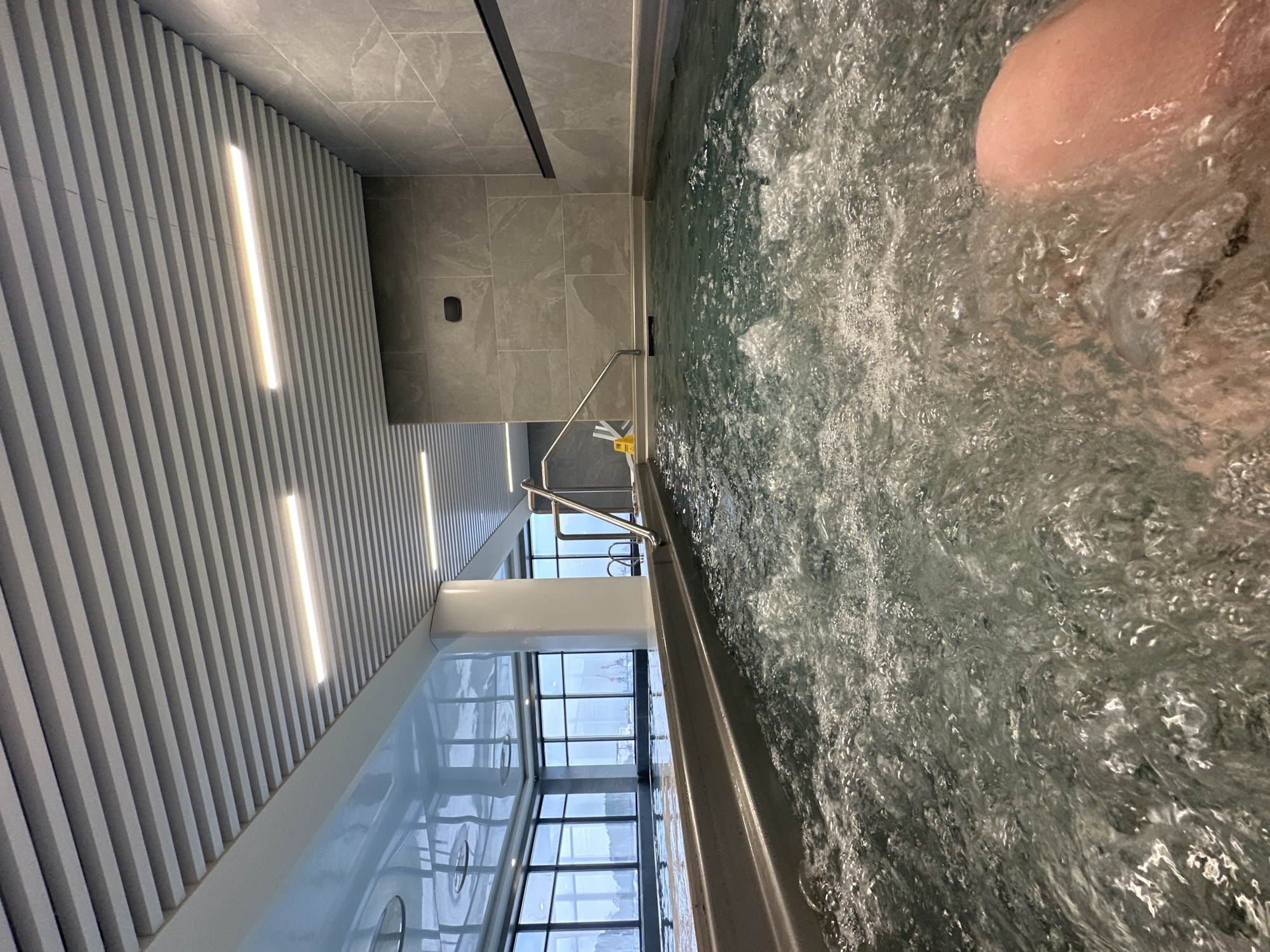 The image shows a first-person view from inside a hot tub, with rippling water filling most of the frame. A portion of a human leg is visible in the upper right corner. The hot tub is located in a modern-looking room with grey-toned walls and ceiling. A section of the ceiling has linear LED lighting integrated into a ribbed design. A stainless steel grab rail is visible near the edge of the tub. Through a large window in the distance, a cityscape can be observed, suggesting the location is in an urban area. While the prompt specifies Chaikovsky, Russia, nothing within the image specifically confirms this location.