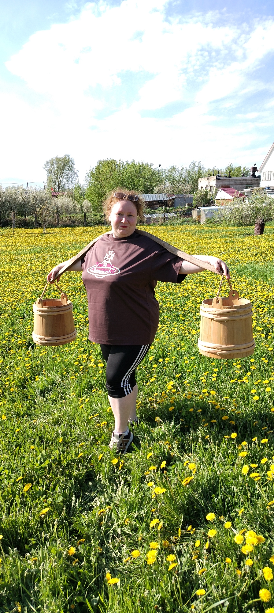 A woman stands in a field of dandelions in what appears to be Chaikovsky, Russia, holding two wooden buckets suspended from a yoke. She is wearing a brown t-shirt with a logo, black leggings with white stripes, and athletic shoes. The backdrop includes a mix of houses, trees, and blossoming shrubs, suggesting a rural or suburban area. The sky is clear and bright.