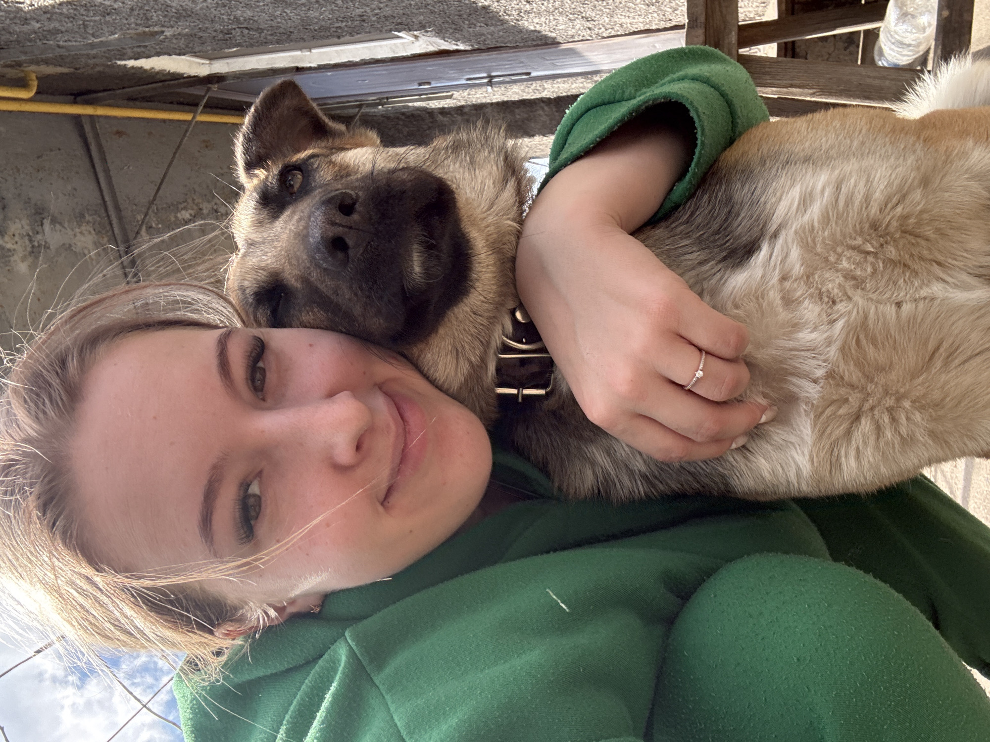 A woman is holding a dog in an outdoor location. The woman is wearing a green hoodie and has light-colored hair. She is smiling at the camera. The dog is brown and tan, with a black collar. The woman's hand is visible with a ring on her ring finger. The background includes a building with a yellow pipe, a wooden structure, and a water bottle. The exact location within Donetsk, Ukraine, cannot be determined from the image.